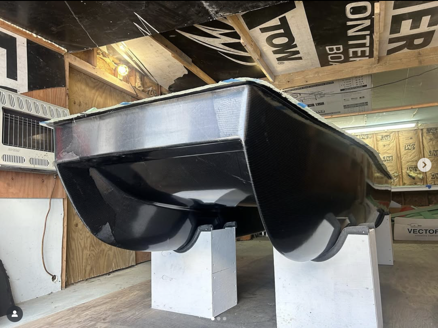 A carbon fiber boat  in an unfinished garage or workshop, resting on two white stands with wooden walls and insulation in the background.