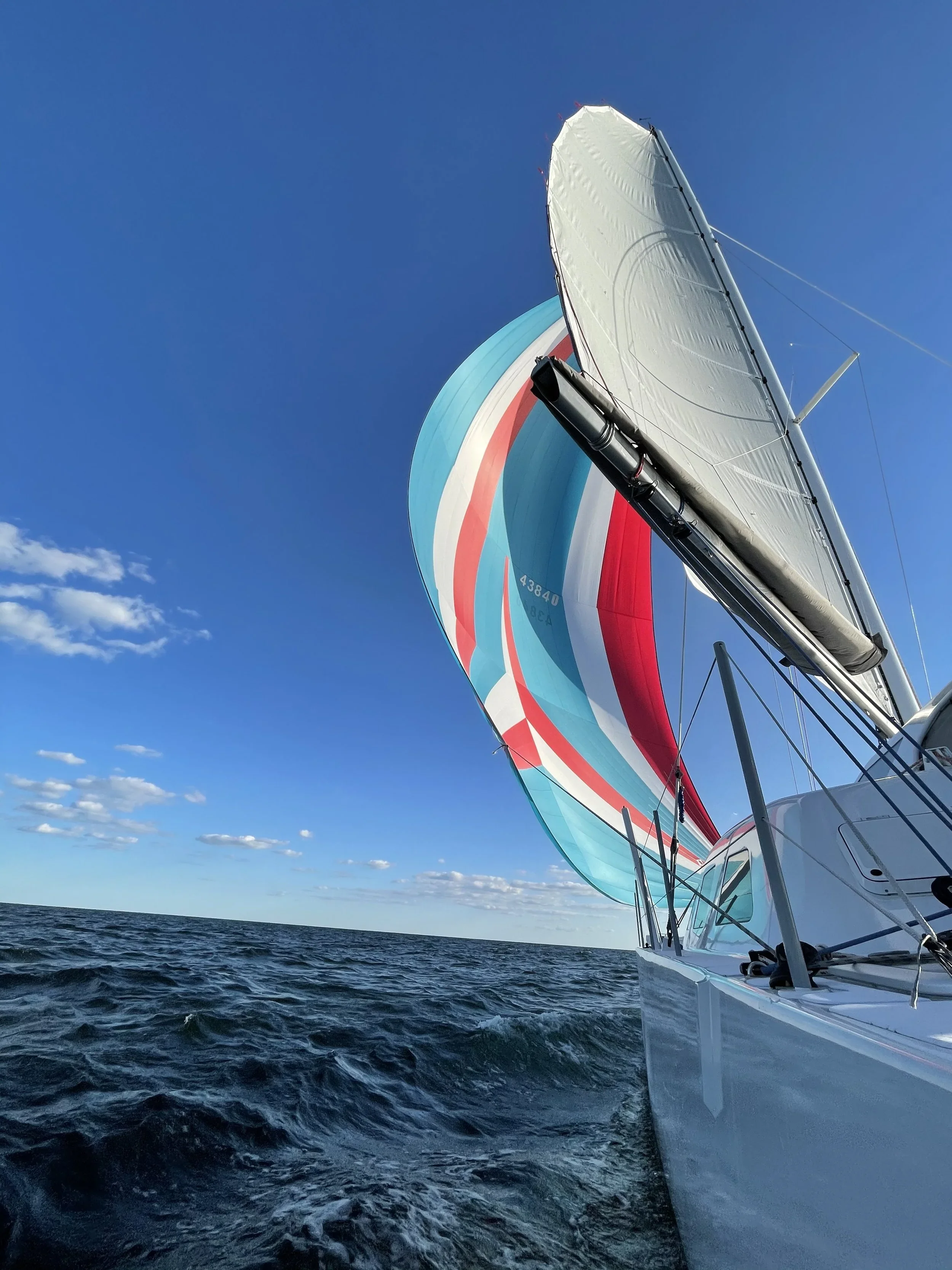 View from a sailboat on the open ocean during daytime, showing the boat's sail with red, white, and blue stripes, and the deep blue sky with a few clouds.