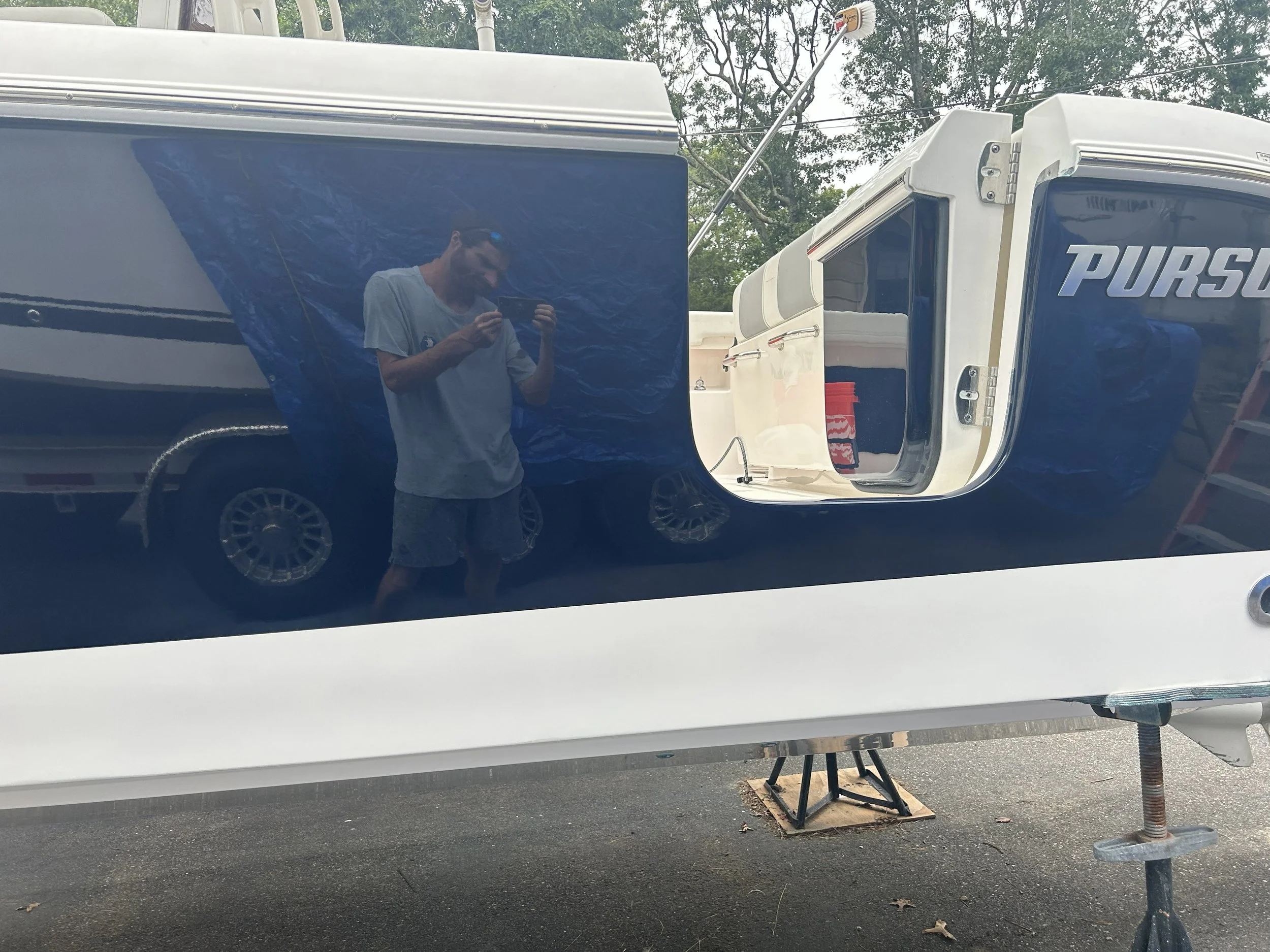 A reflected image of a man taking a photo in the shiny surface of a large black van or trailer. The man is wearing glasses, a light-colored T-shirt, and is smiling. The background includes trees and a cloudy sky, with some equipment and support blocks under the vehicle.