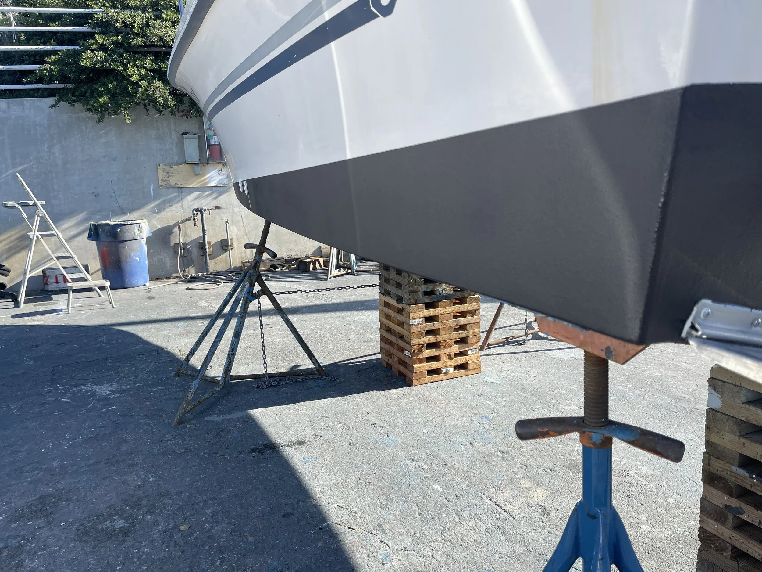 View of an outdoor boat workshop with a boat on jack stands, wooden pallets supporting the boat, tools, a ladder, and a trash bin visible, and greenery in the background.