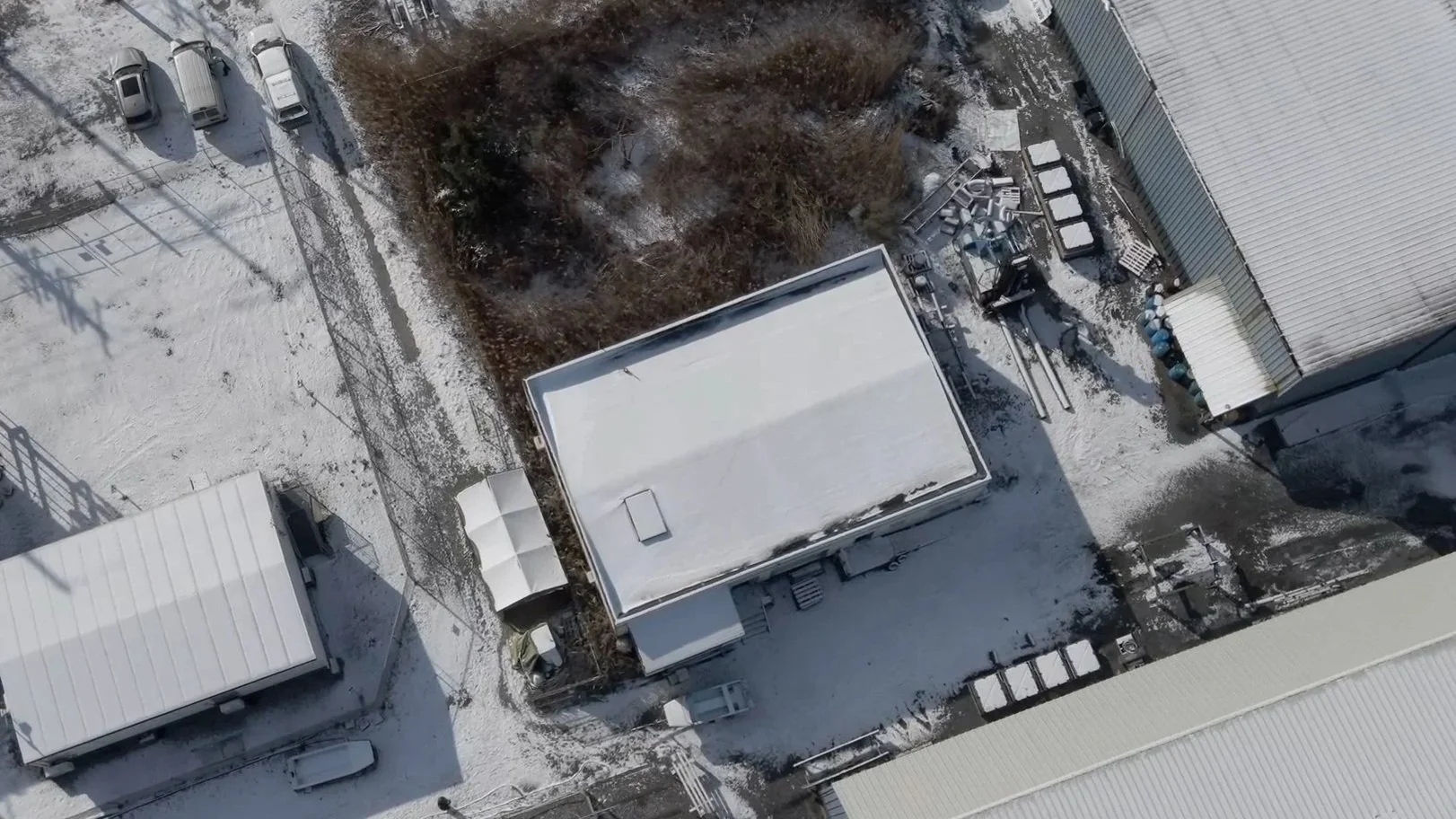 An aerial view of a snow-covered industrial or commercial area with multiple buildings, parked cars, and scattered outdoor equipment, with some trees and a dirt path visible.