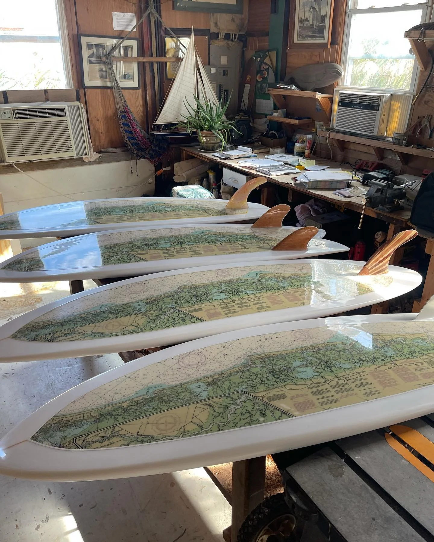 Several white paddleboards with topographical and navigational maps on their surfaces are arranged in a workshop, with a cluttered workbench, window air conditioning units, framed pictures, and various tools in the background.
