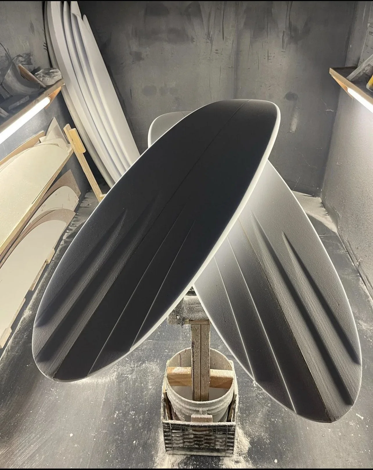 A surfboard with a black top and white edges on a wooden stand inside a workshop, with other surfboards leaning against the wall.