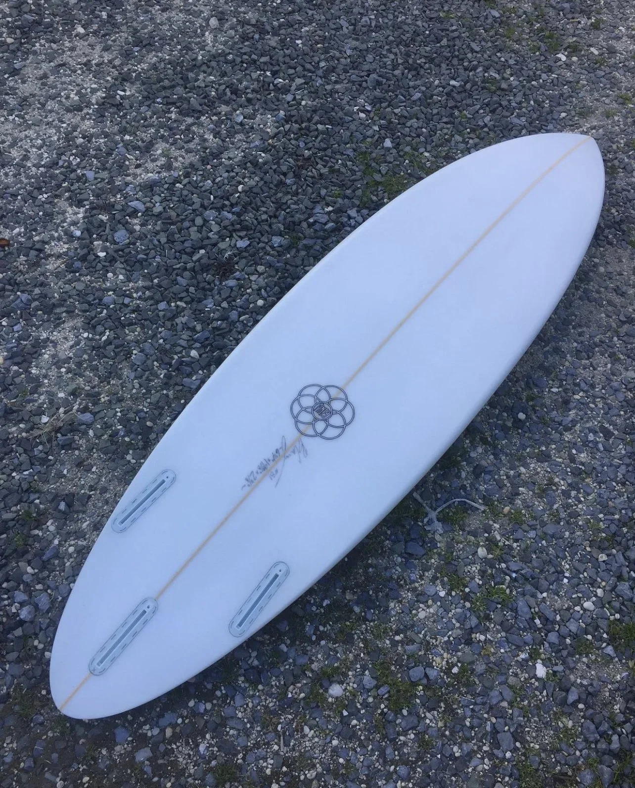 A white surfboard lying on gravel with a yellow string running down the center and a metal logo in the shape of a flower near the center.