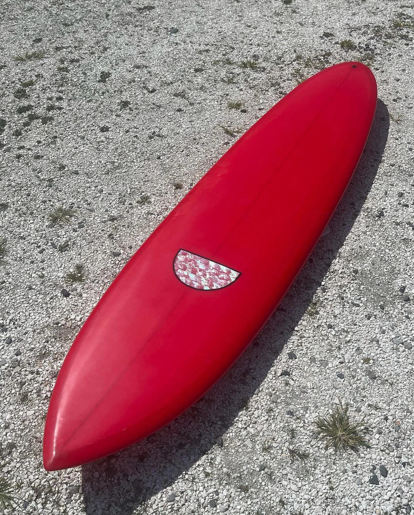 Red surfboard lying on a gravel surface with small plants, casting a shadow.