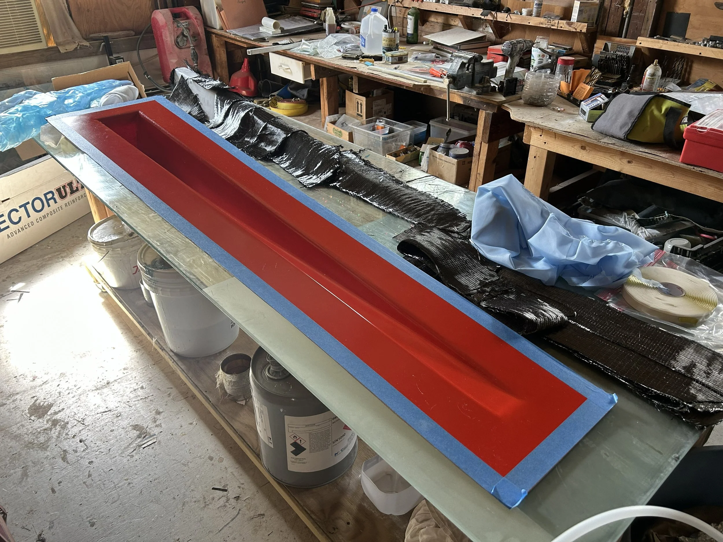 A cluttered woodworking or workshop space with a workbench filled with tools, supplies, and equipment. There is a long, rectangular object made for composite tooling- to be used as a mold for vacuum bagging and vacuum infusion.