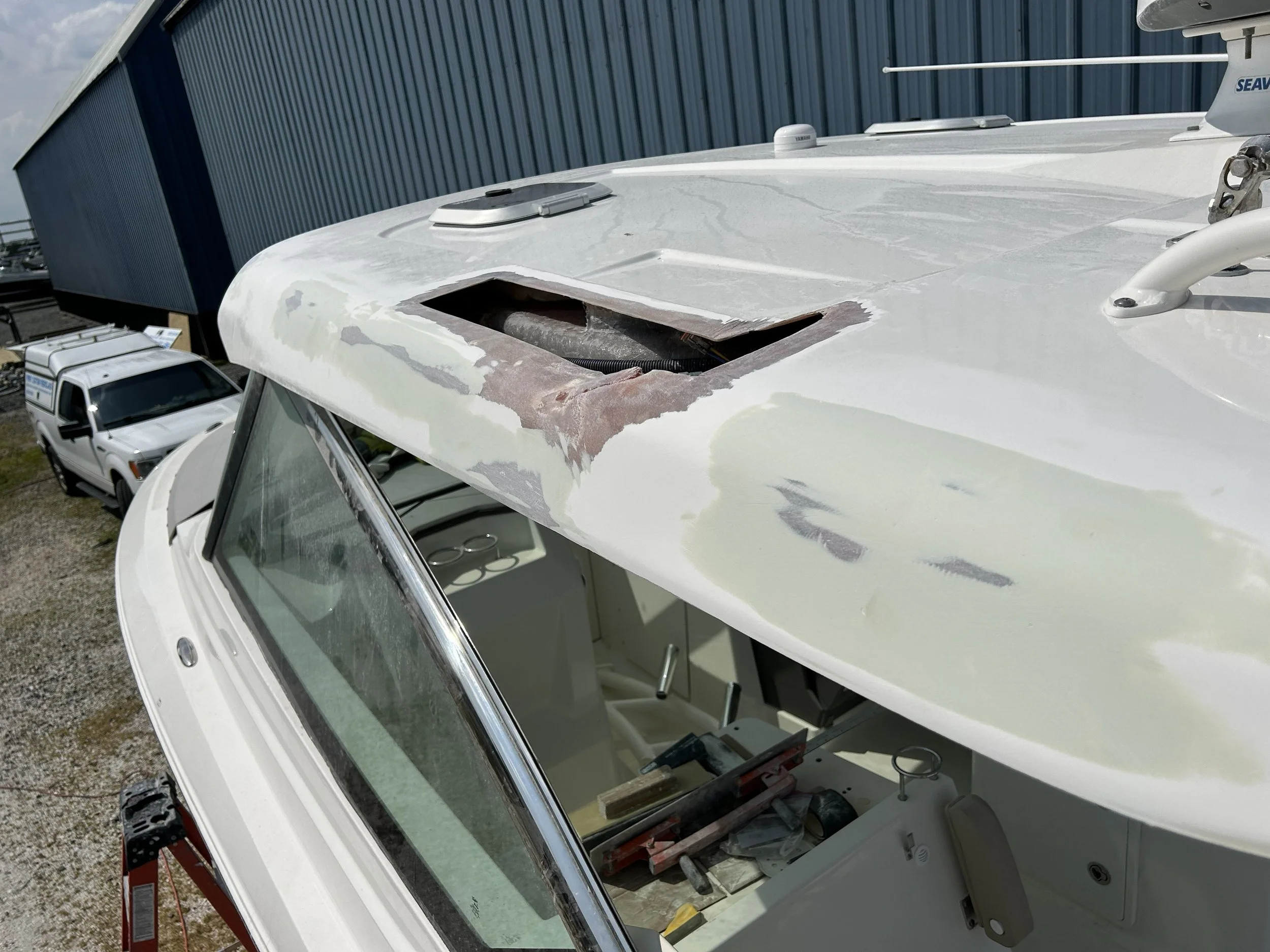 The image shows the top of a white RV or camper with a damaged or removed roof vent, exposing the internal structure. There is a window with a reflection of the sky on the front side of the RV, with tools and materials nearby, and a blue building in the background.