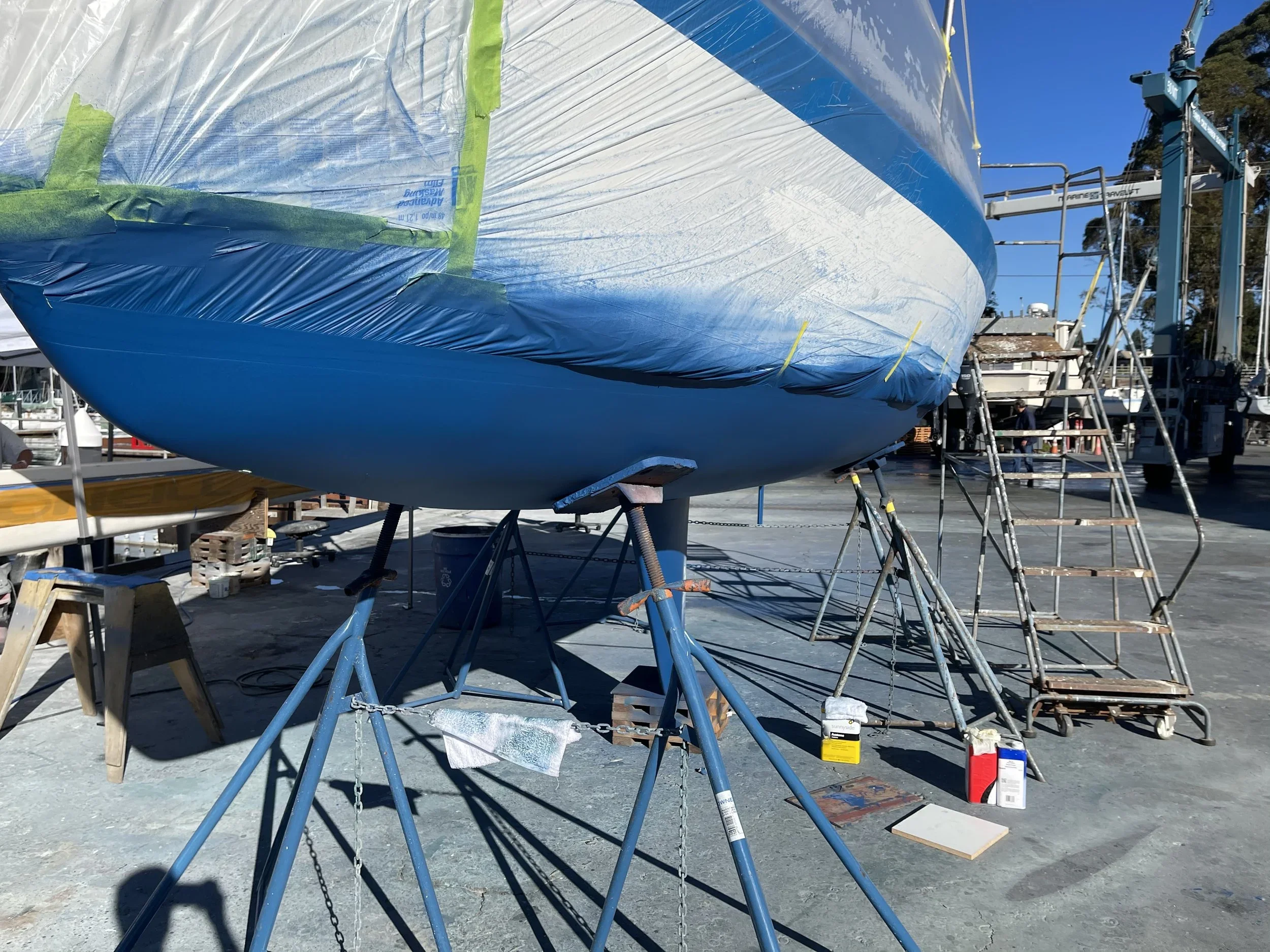 A boat is being repaired or built outdoors, covered in blue and white protective wrapping, supported by blue stands, with ladders, tools, and supplies around it, against a background of clear blue sky and trees.