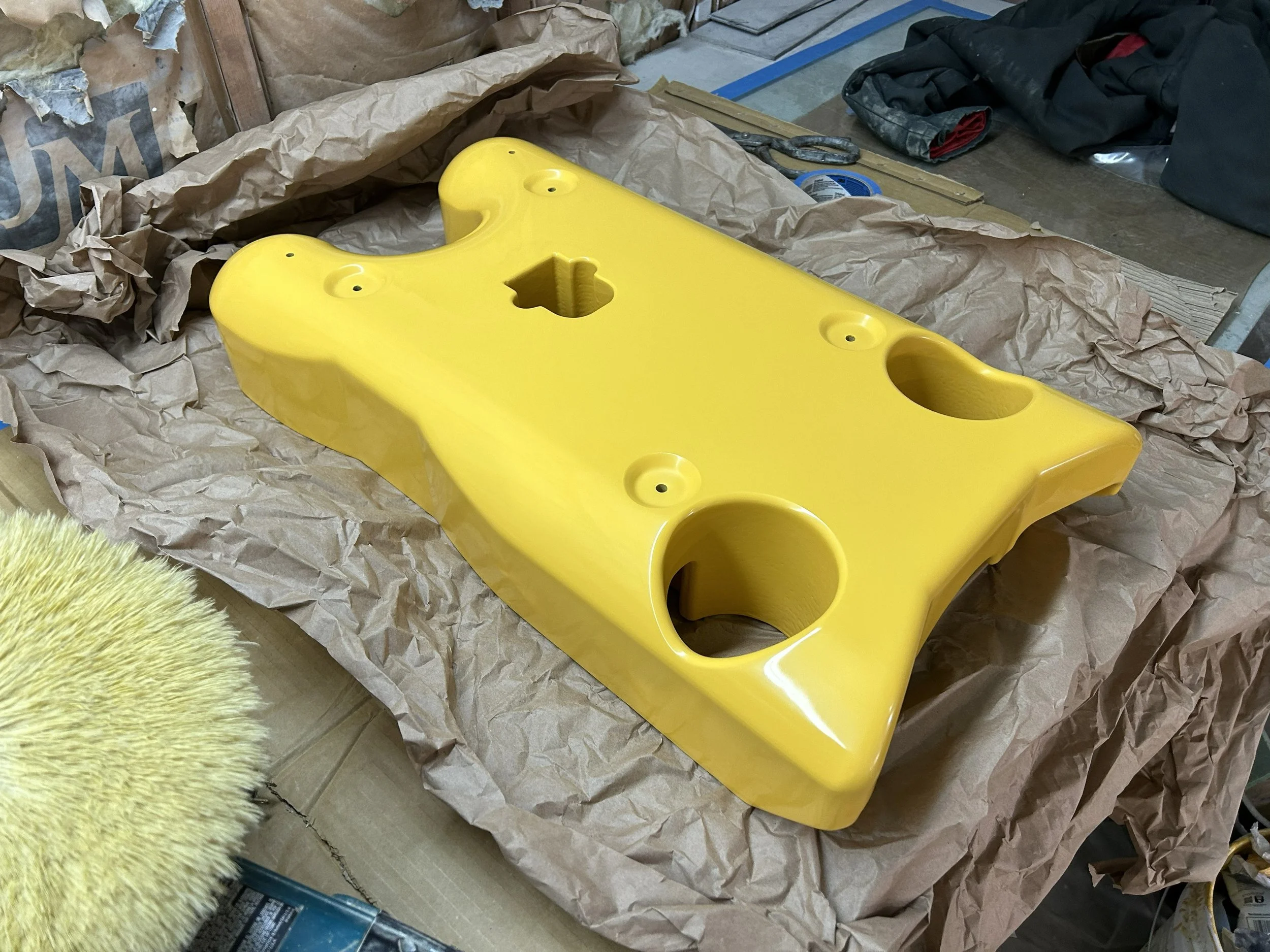 A yellow plastic object with multiple circular openings and a keyhole-shaped opening in the center, placed on crumpled brown paper in a workshop or construction area, with various tools and supplies around.
