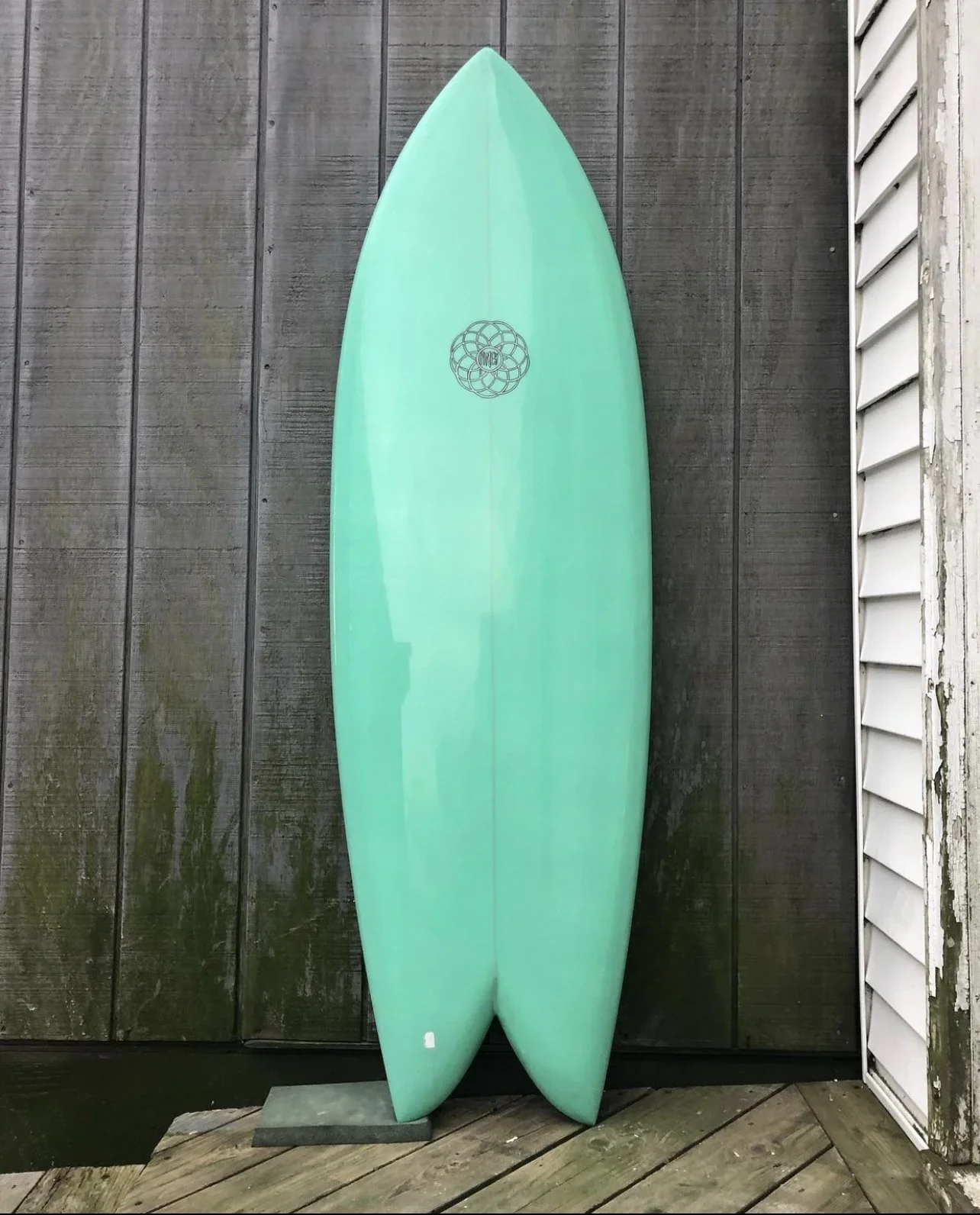Surfboard standing upright against a dark wooden fence, with a white house siding on the right side of the image.