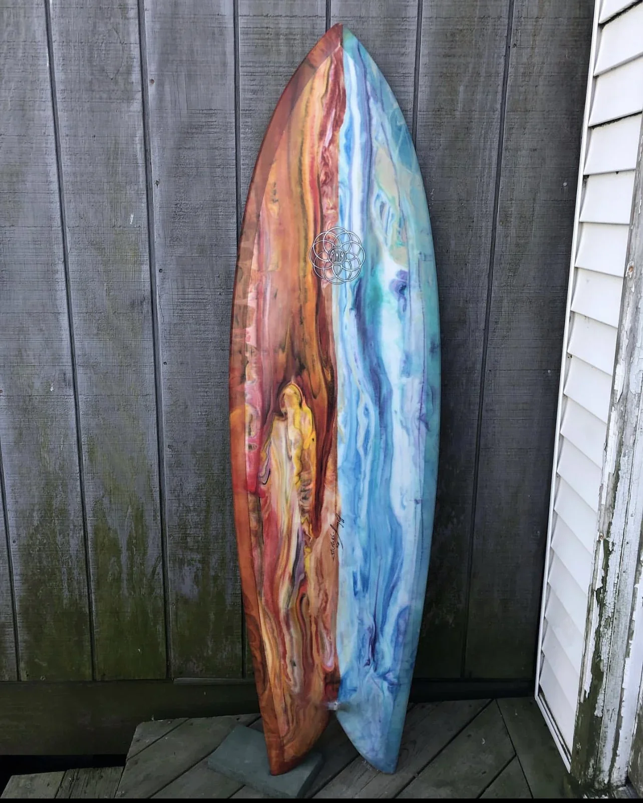 Colorful wooden surfboard with abstract painting design, leaning against a wooden fence outdoors.