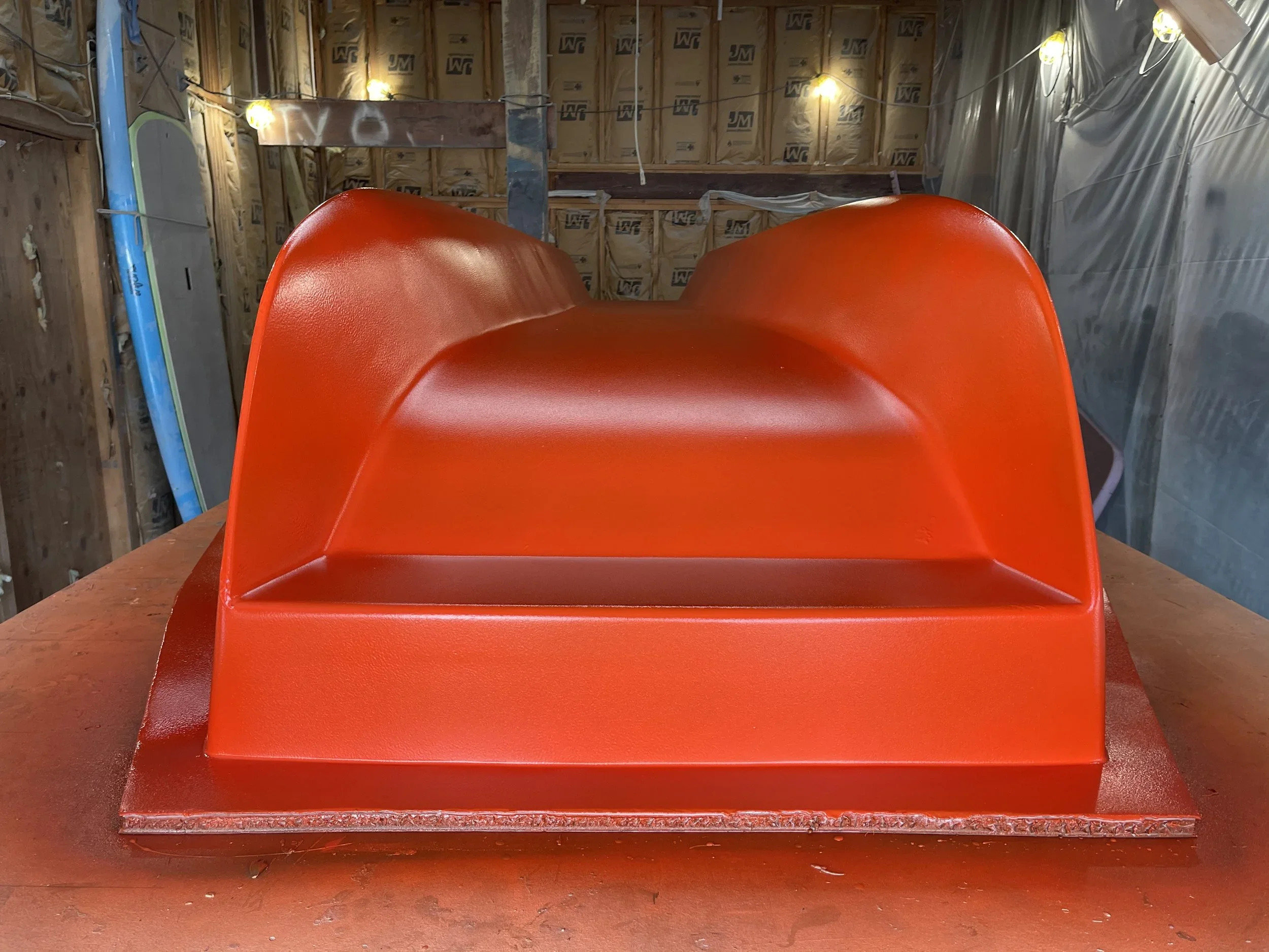 A catamaran dinghy boat mold being built off tooling plug in a partially constructed room with exposed insulation and framing, and a red plastic slide or scoop in the foreground placed on a wooden surface.
