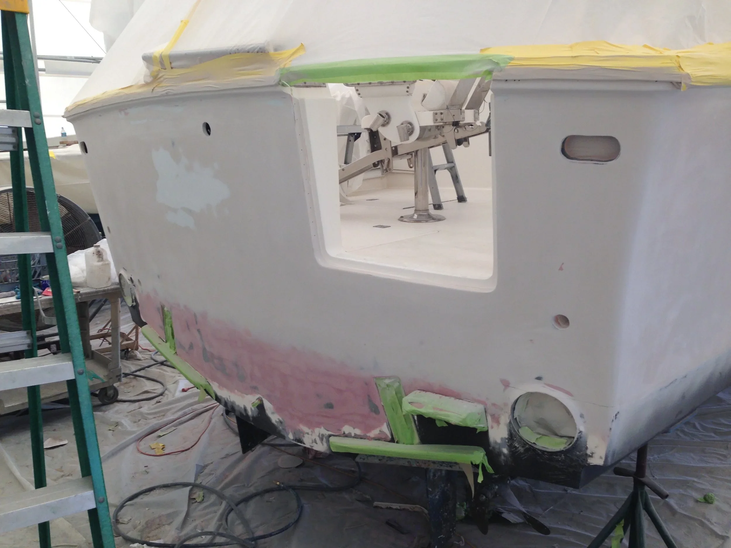 A white boat hull under construction with masking tape and sanding patches, surrounded by scaffolding and tools in a workshop.