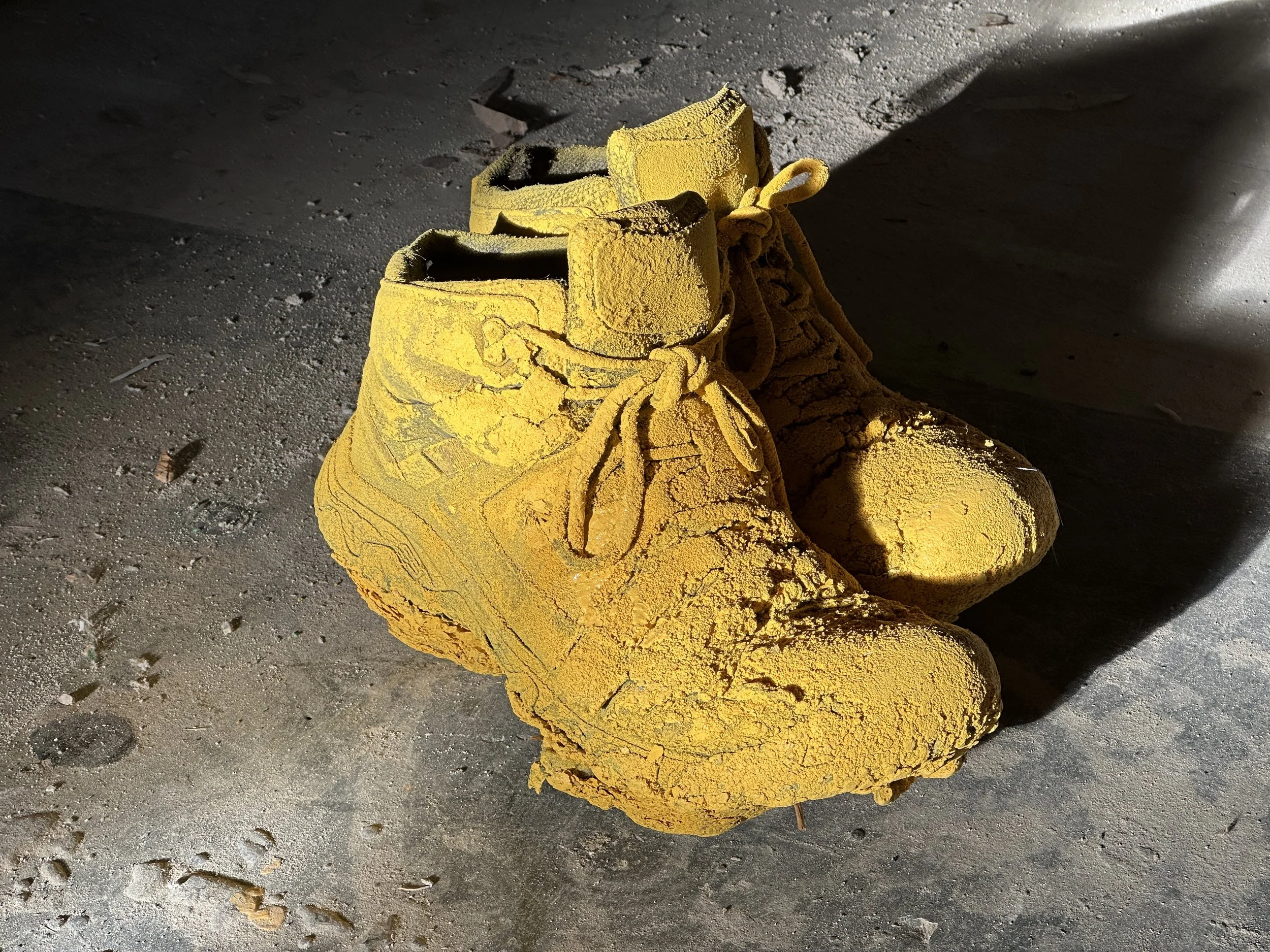 A pair of yellow, dusty hiking boots on a gravel surface, with sunlight casting shadows.