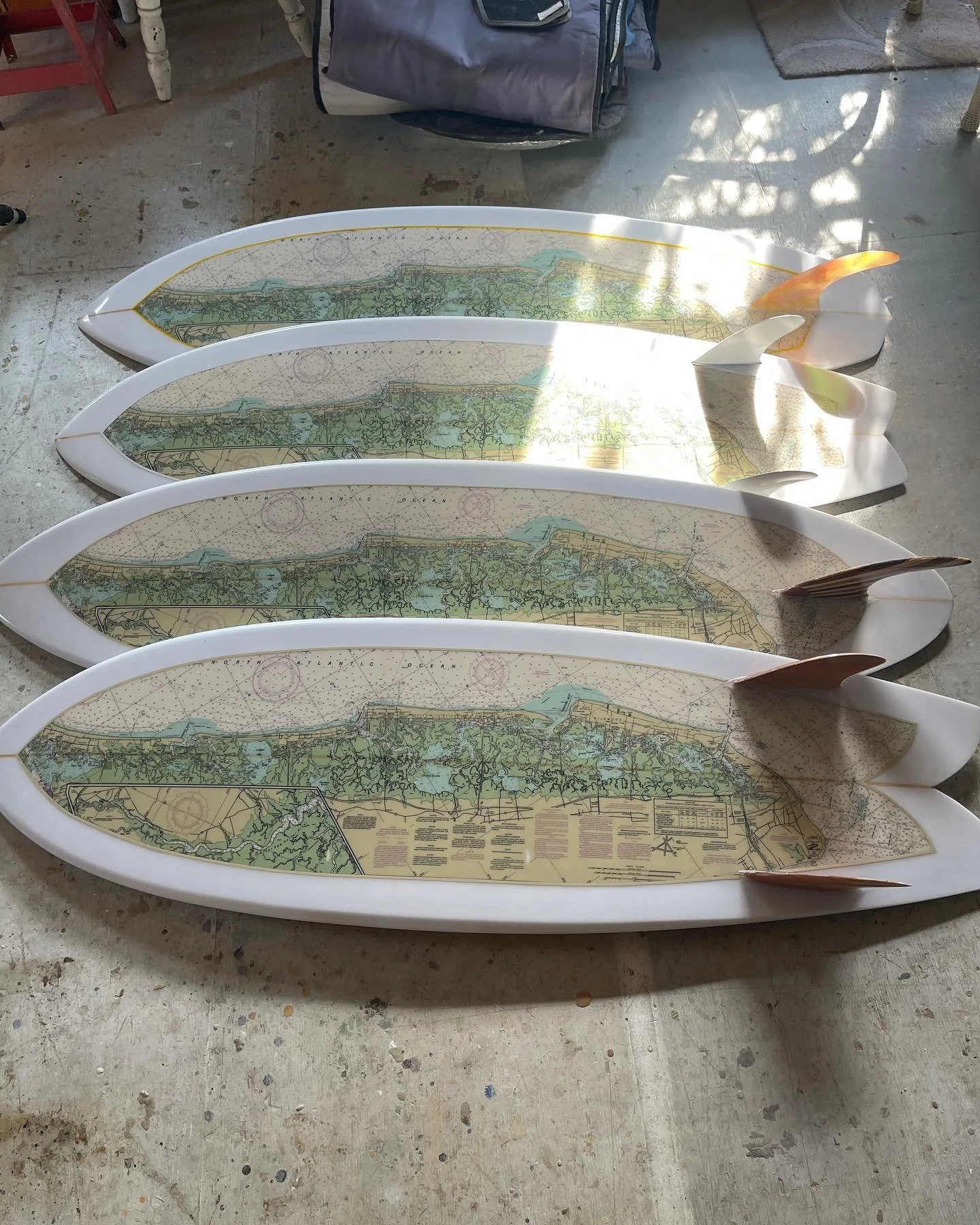 Four surfboard-shaped maps laid out on a concrete floor, with a backpack and chairs nearby, and sunlight creating shadows on the floor.