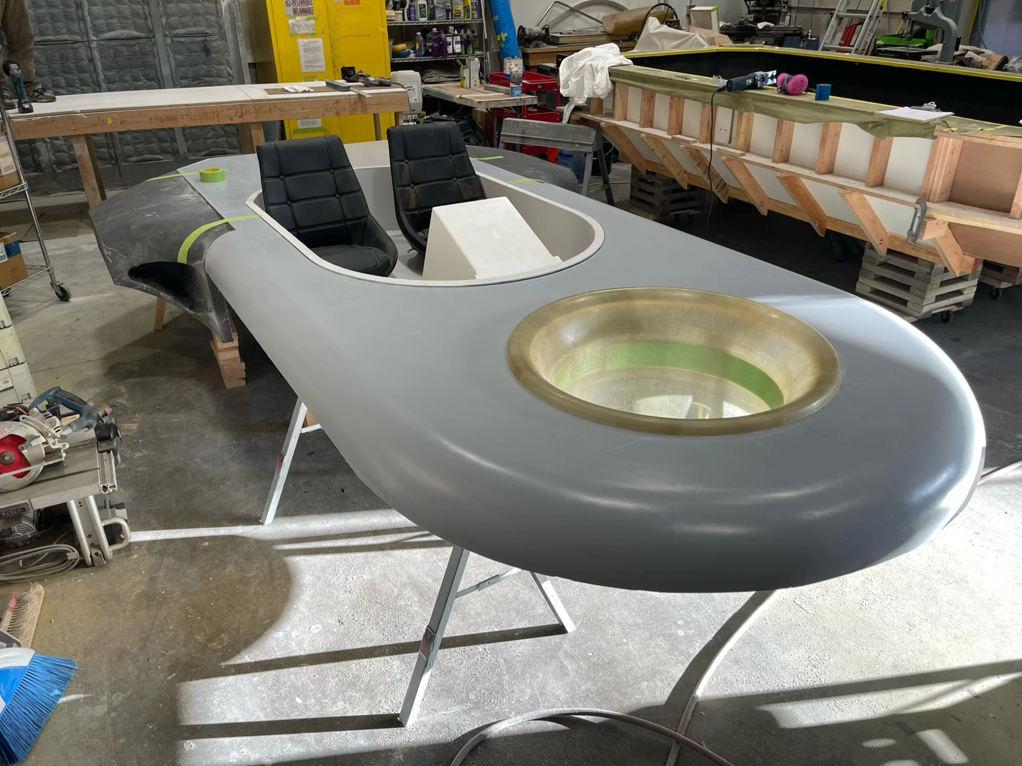 A workshop with a hovercraft under construction, black chairs, tools, shelves, and workbenches.
