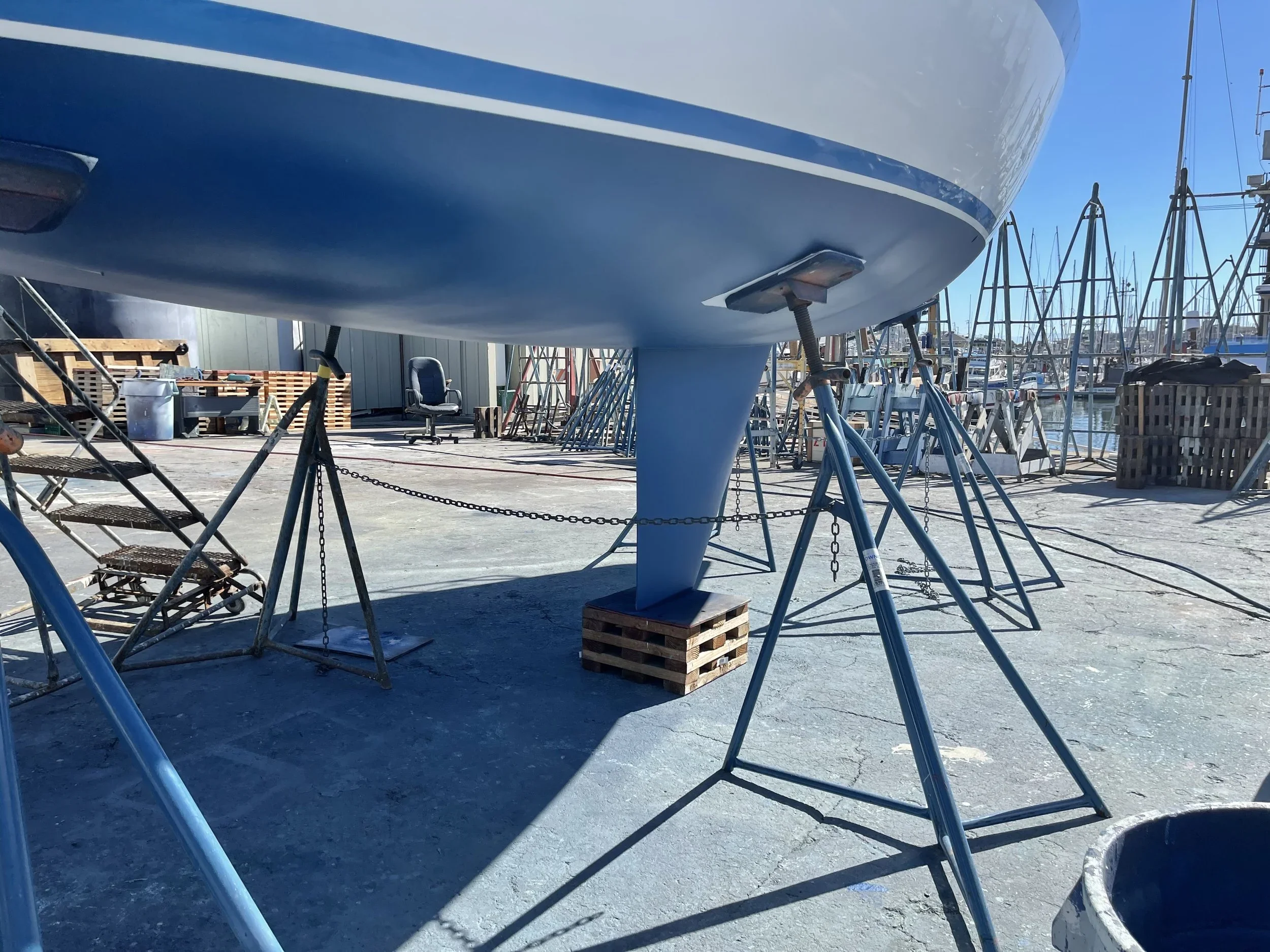 View of a boat hull supported on stands at a marina, with other boats and masts visible in the background.