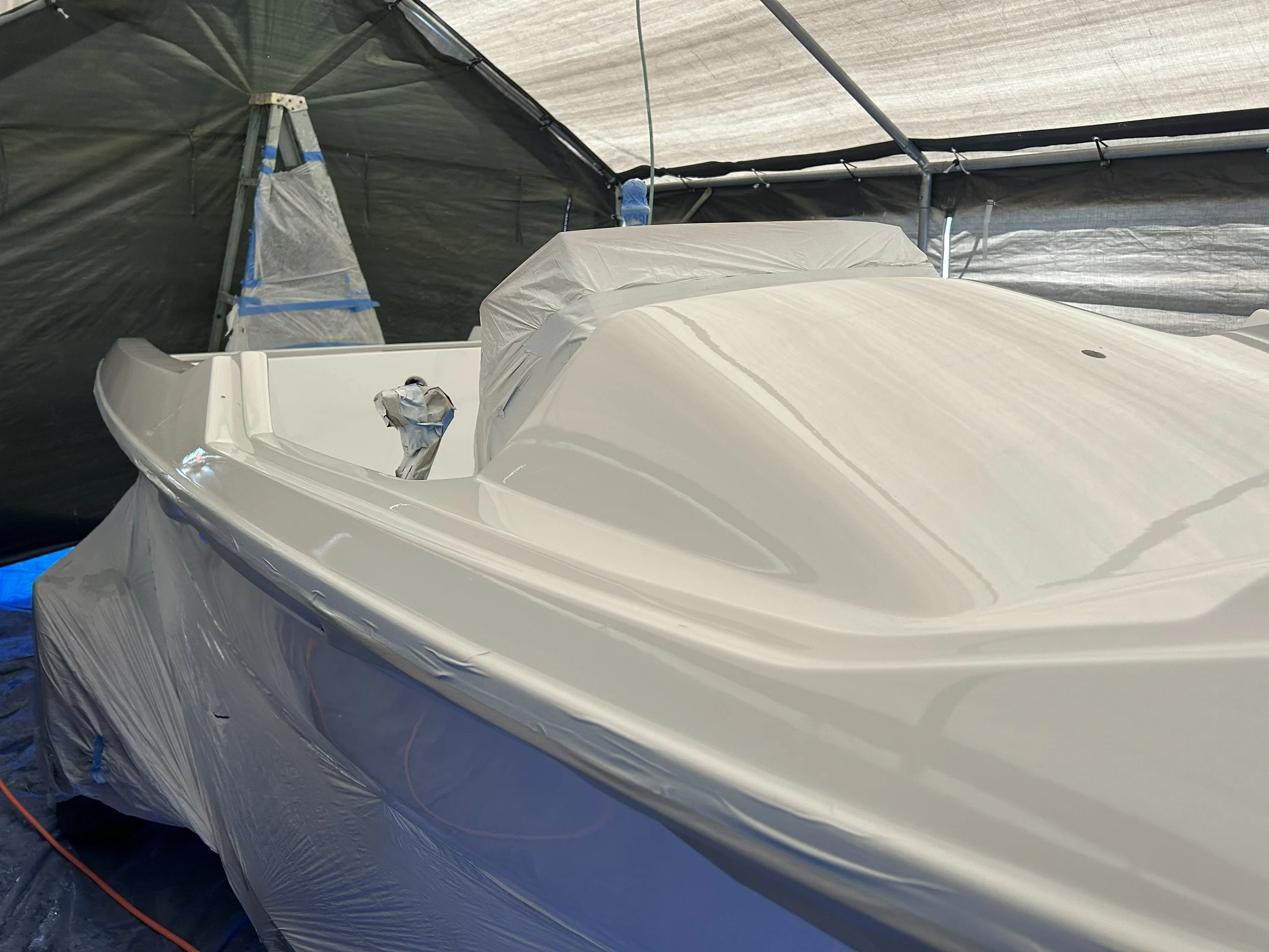 A white boat covered with plastic, inside a large industrial tent with a mostly gray fabric roof, supported by metal poles. The boat and surrounding area appear to be protected or prepared for painting or maintenance. Awlgrip boat paint job.