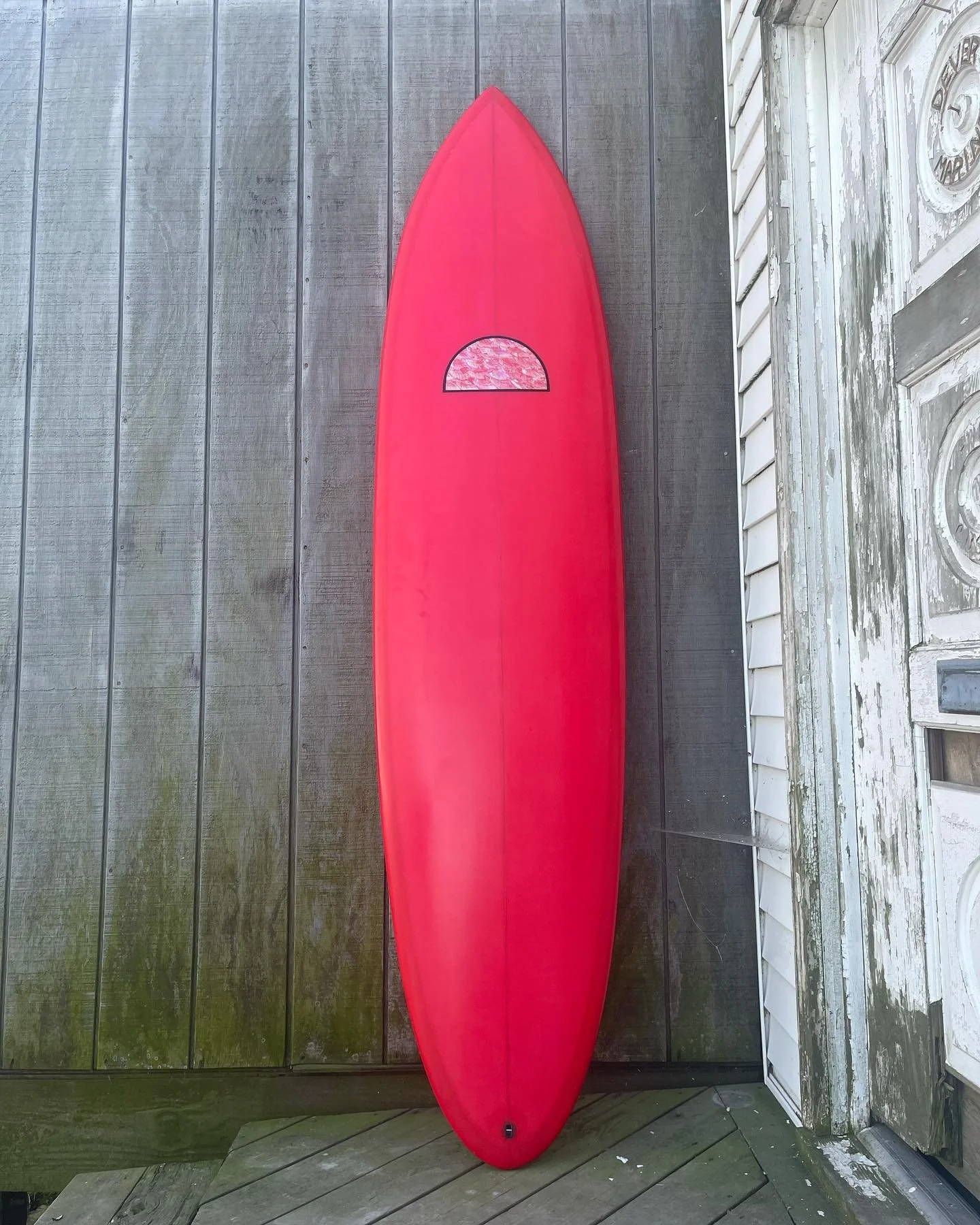 Red stand-up paddleboard with a small window on its front, leaning against a weathered wooden wall on a porch or deck.