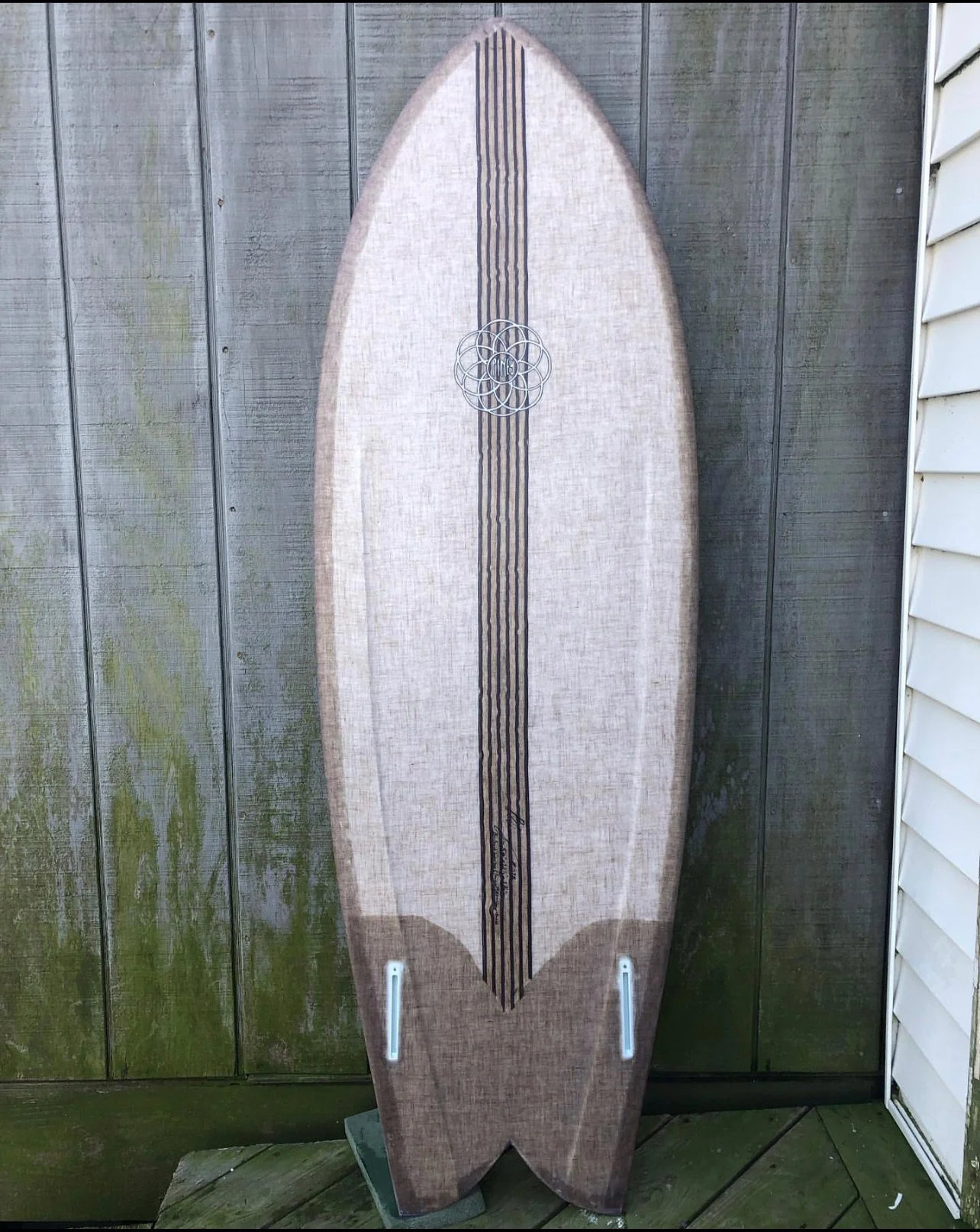 A longboard surfboard standing upright against a wooden fence with moss and algae.