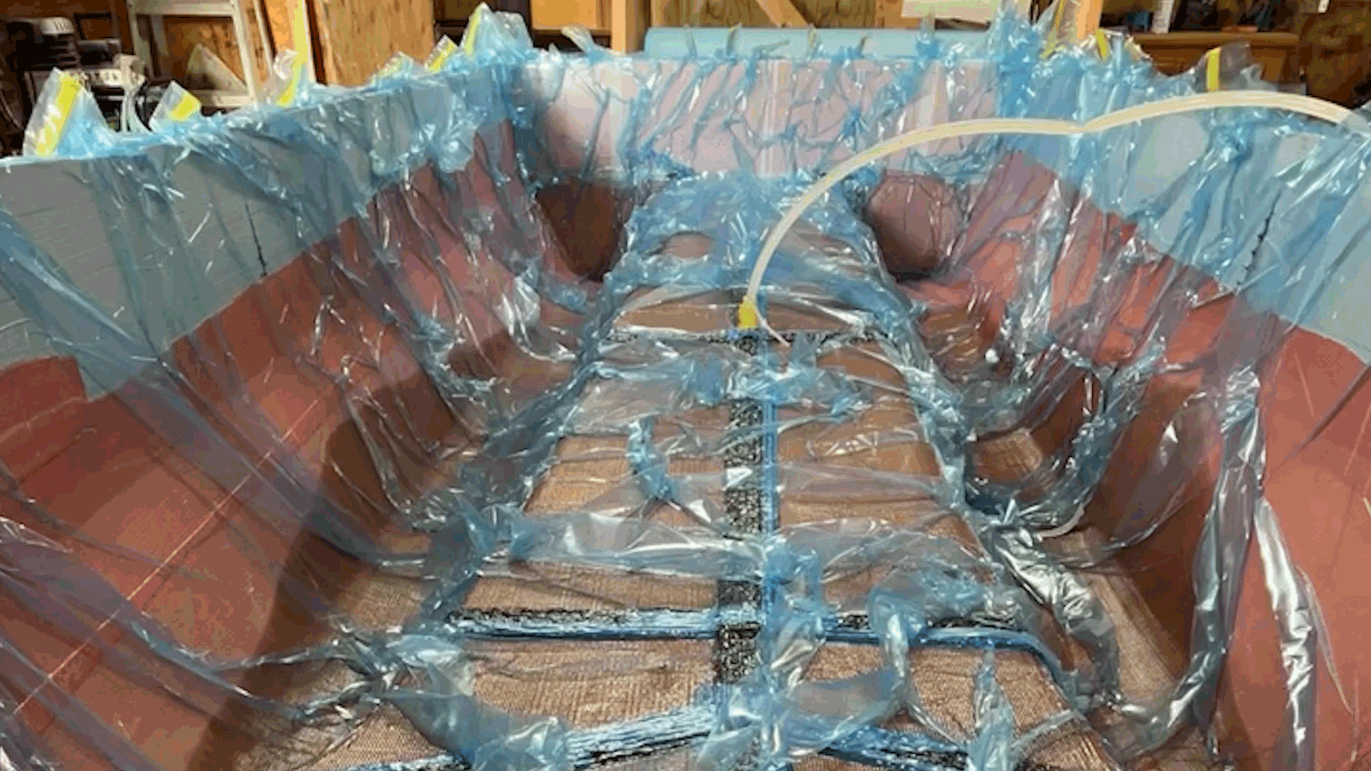 A boat mold is covered with a clear plastic sheet and blue painter's tape, likely in the process of being assembled or protected in a workshop or garage.