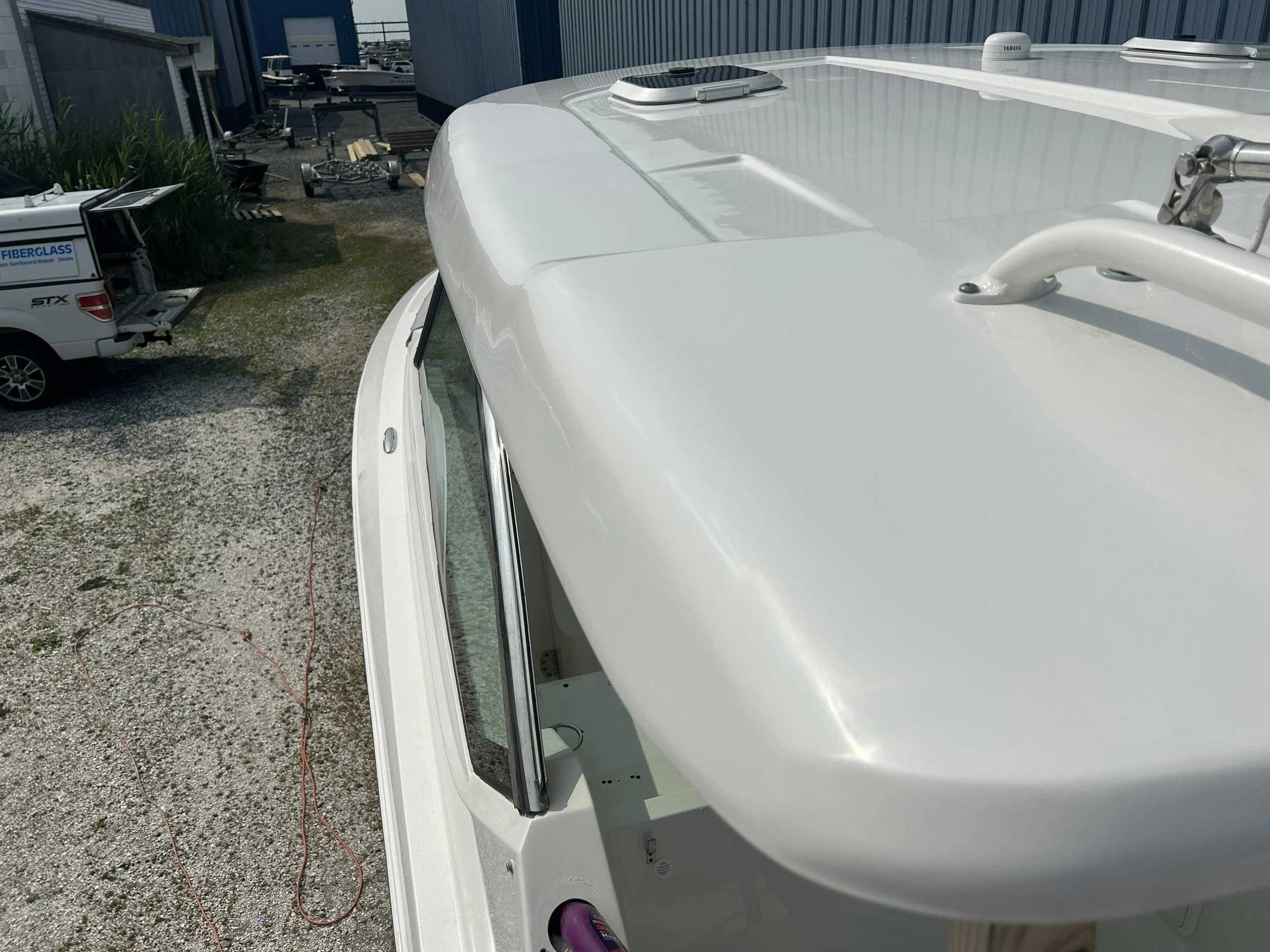 A white boat with a Yamaha rooftop antenna parked outdoors, with other vehicles and boats in the background, and a gravel ground. Finished gelcoat repair on top of boat T top.
