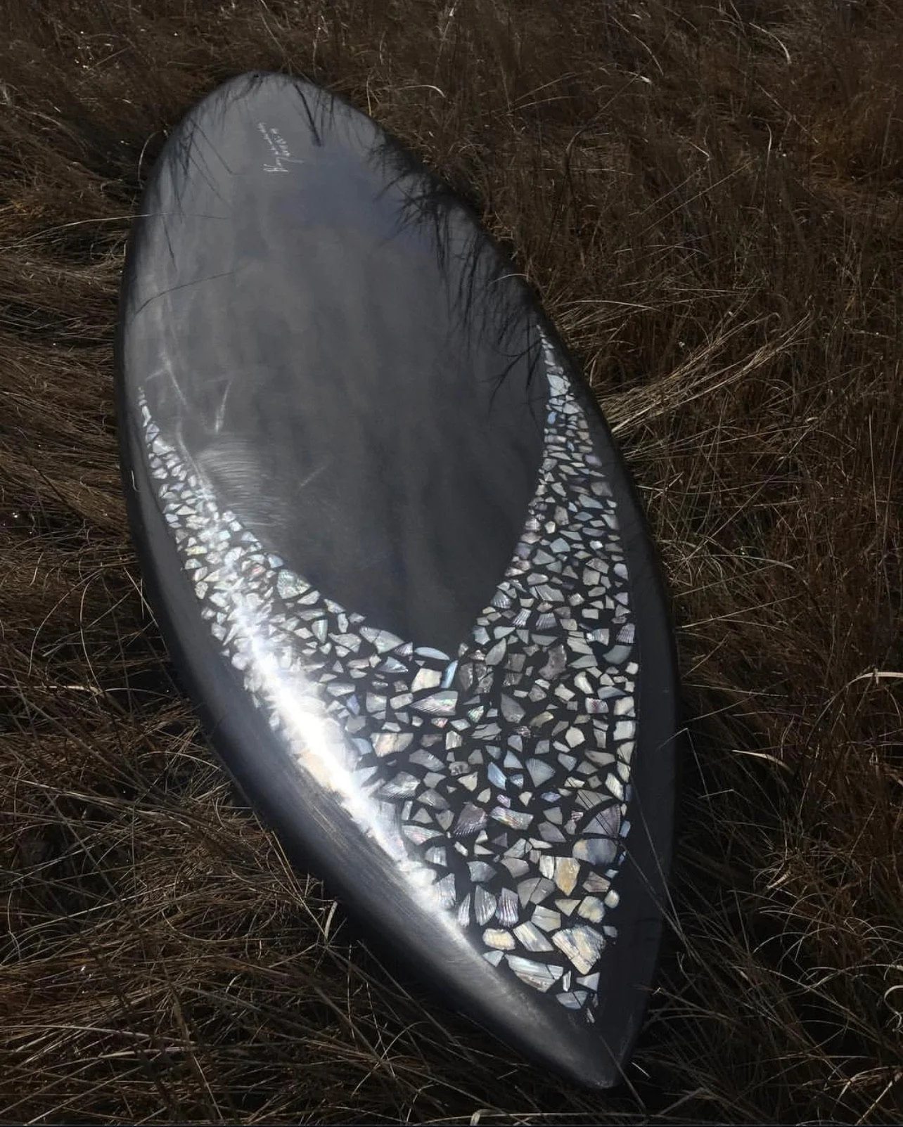 A surfboard with a black border and an iridescent shell mosaic design on the top, lying on dry grass.