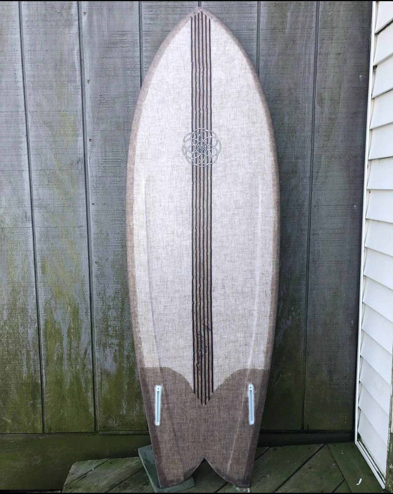A surfboard with a brown stripe pattern, leaningagainst a weathered wooden fence outdoors.