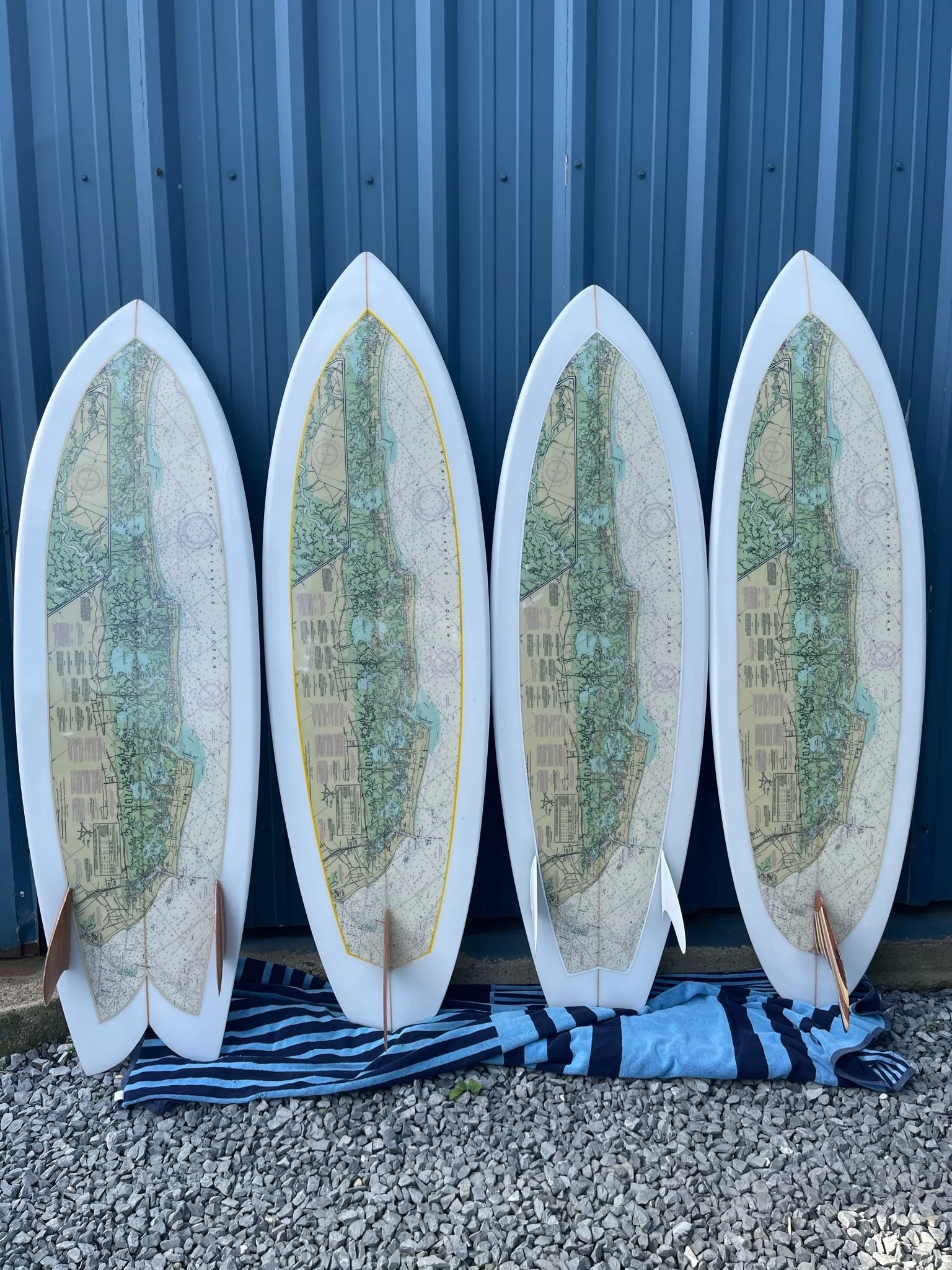 Four surfboards with navigational charts standing upright against a blue corrugated metal wall and resting on a striped fabric on gravel ground.