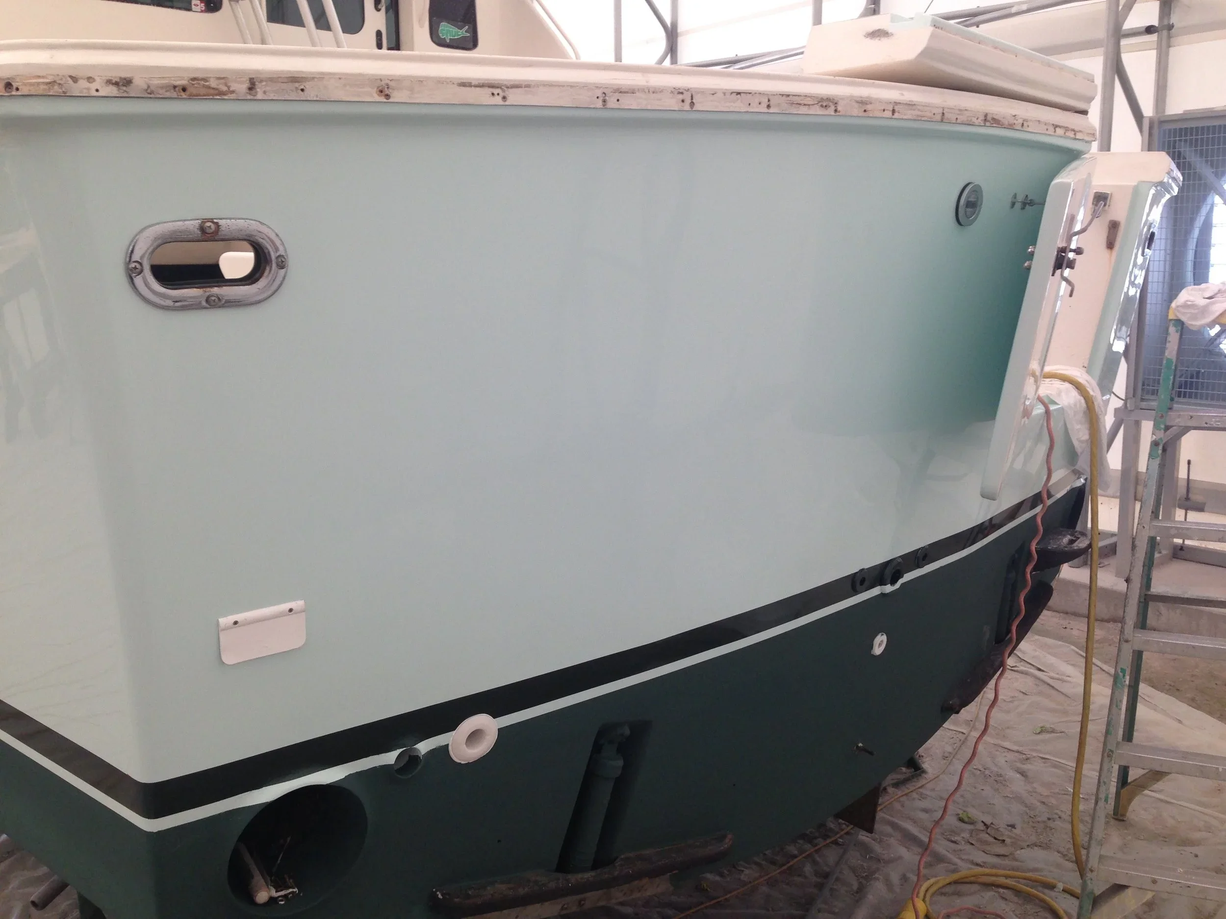 A large boat or yacht in a warehouse or repair facility, with a pale green hull, metal fittings, and a ladder nearby.