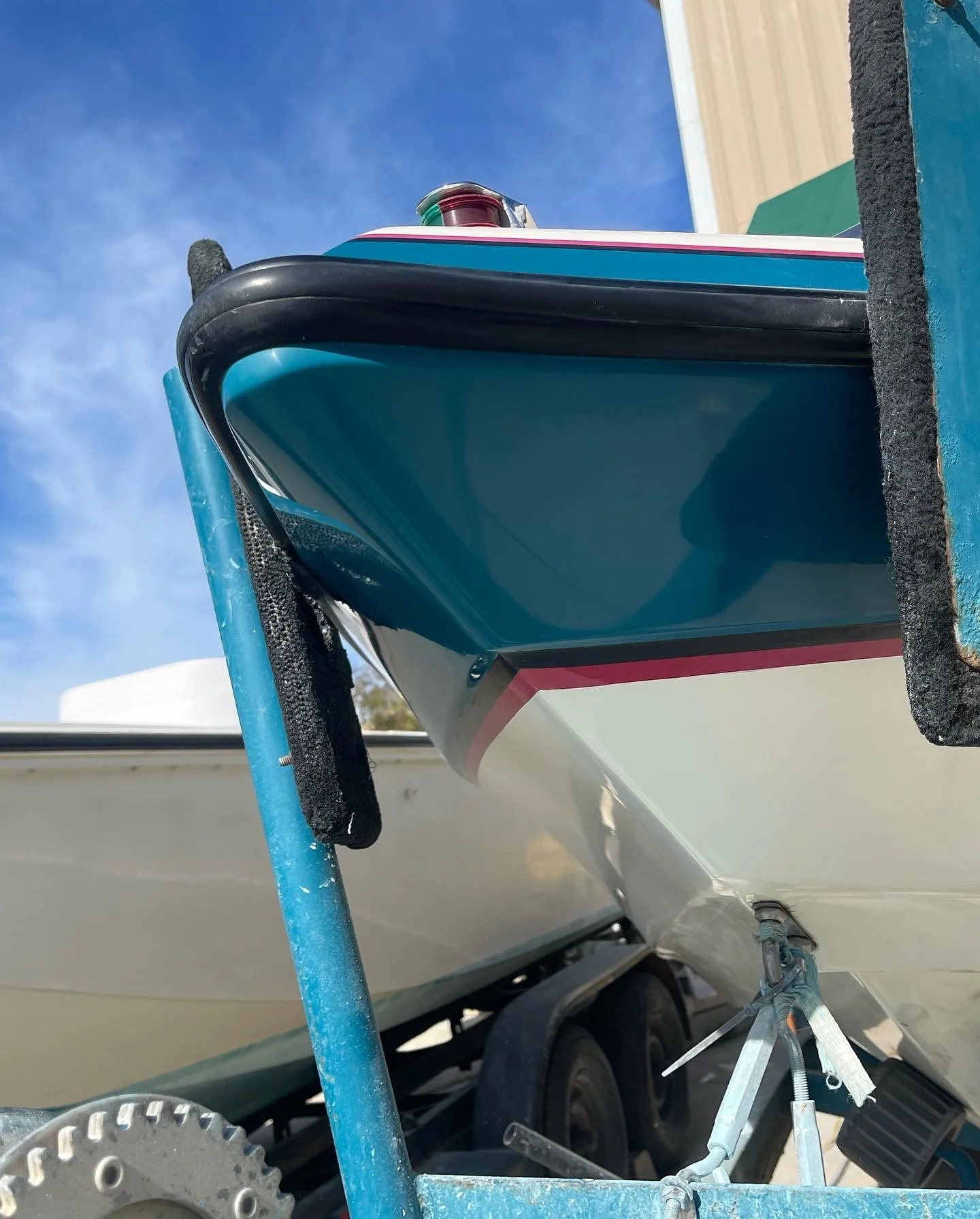 Close-up of a boat hull on a trailer, showing the bow finished matching gelcoat repair with a red and green navigation light, black rubber bumper, and parts of the boat's underside, with a blue sky in the background.
