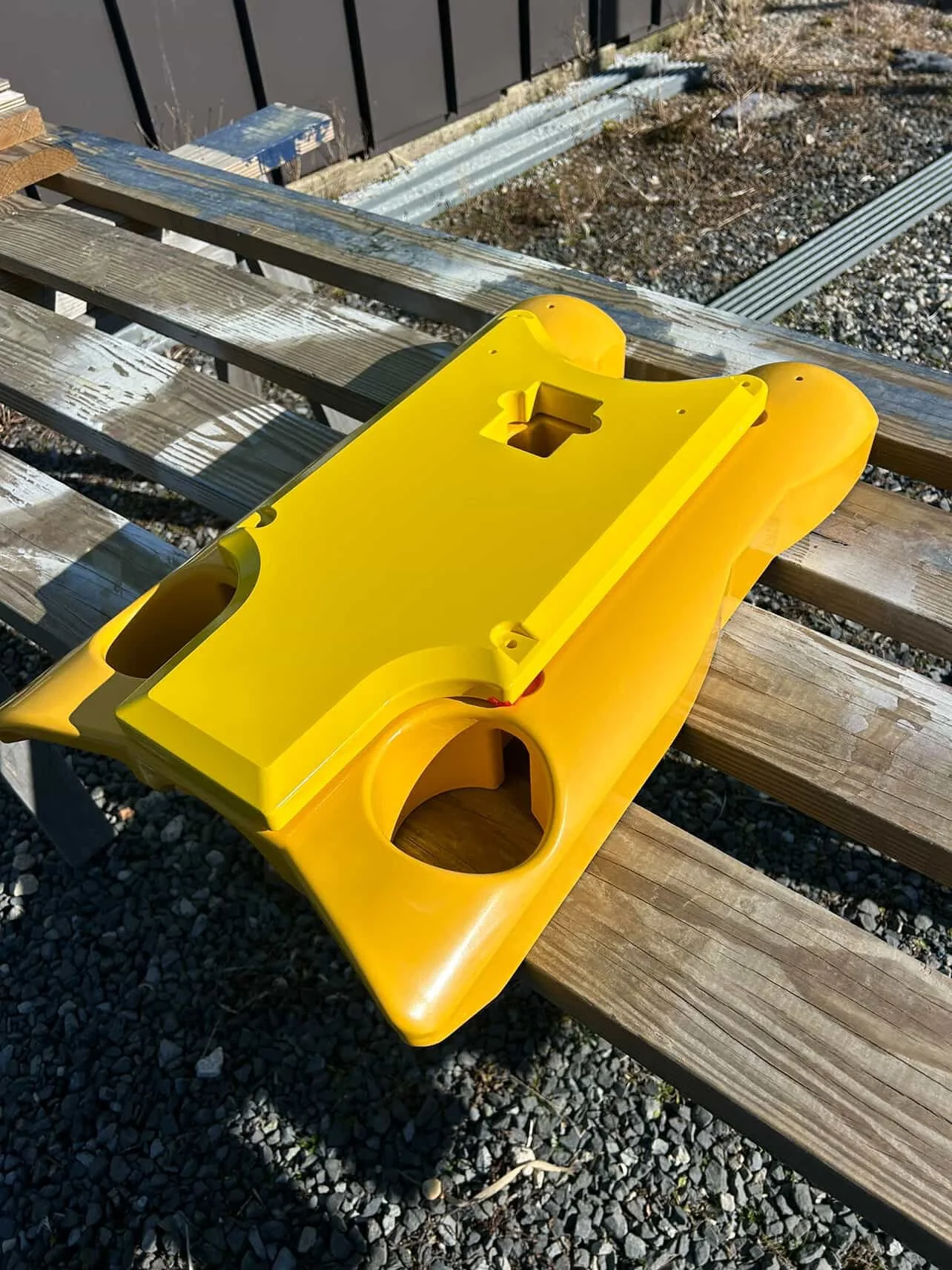 A yellow plastic device with holes and compartments, placed on a wooden bench outdoors, with gravel ground and some metal siding in the background.