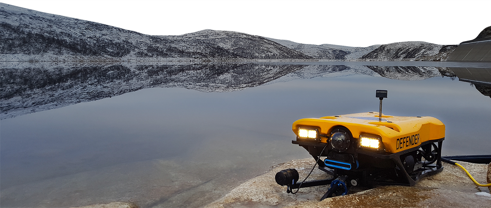 An autonomous yellow underwater drone labeled 'DEFENDER' on a rocky shoreline by a calm lake with snow-covered mountains in the background.