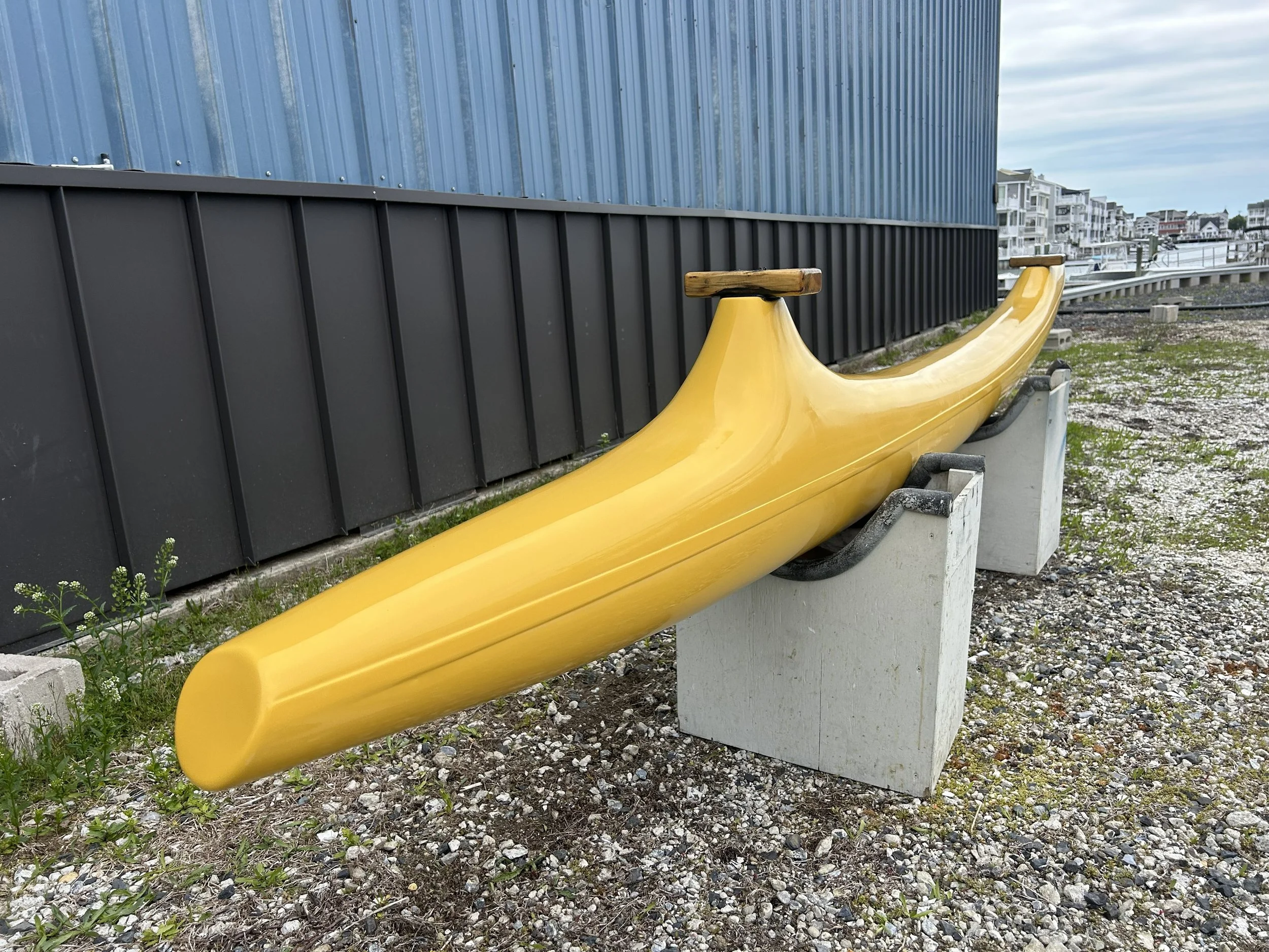 Yellow boat hull used for canoeing positioned outdoors on a gravel surface next to a building with blue siding and a black metal wall. Fresh repaint of gelcoat and wax buff and polish .