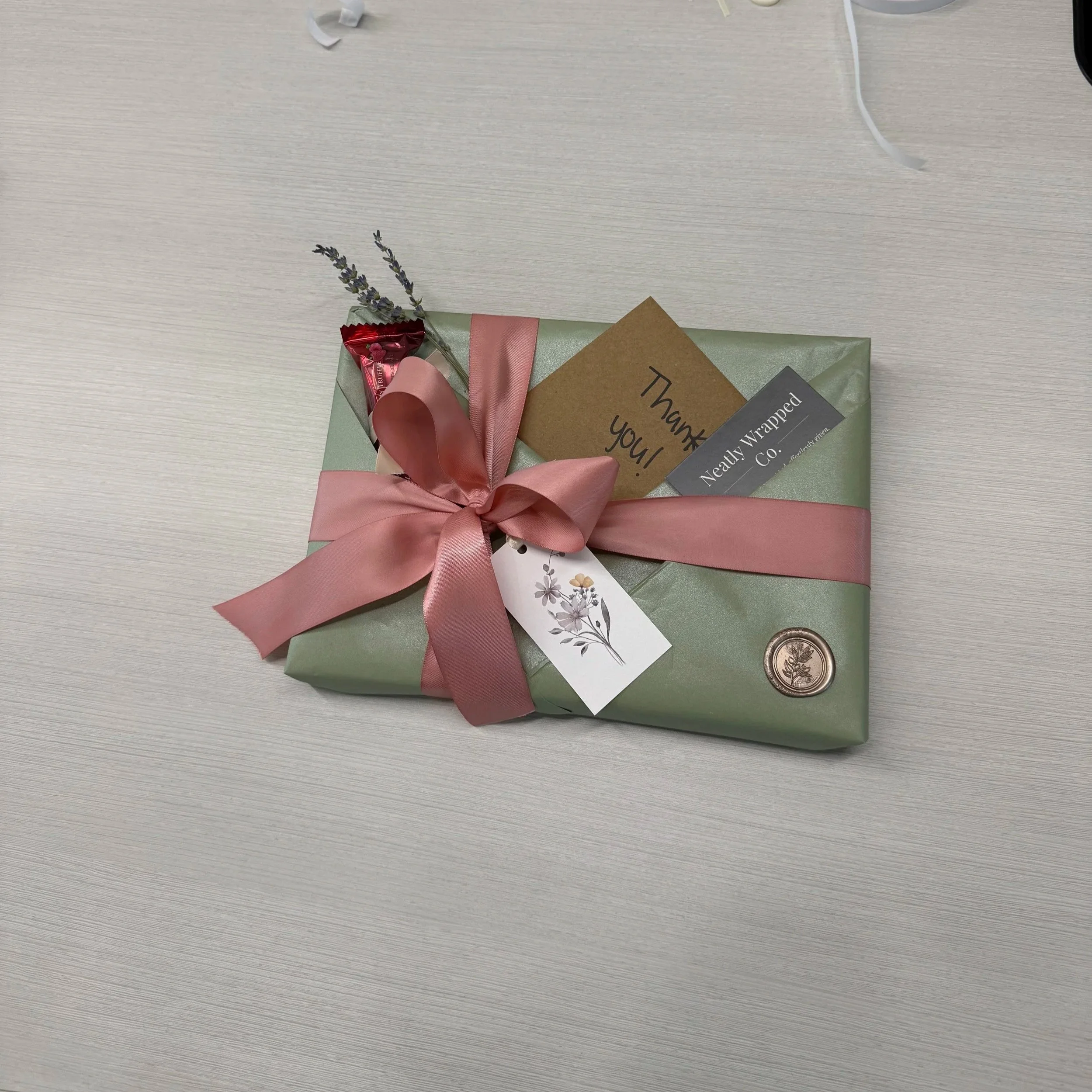 Gift wrapped in light green paper with a pink ribbon, lavender sprig, thank you card, and small chocolate with a gift tag on a light wood surface.