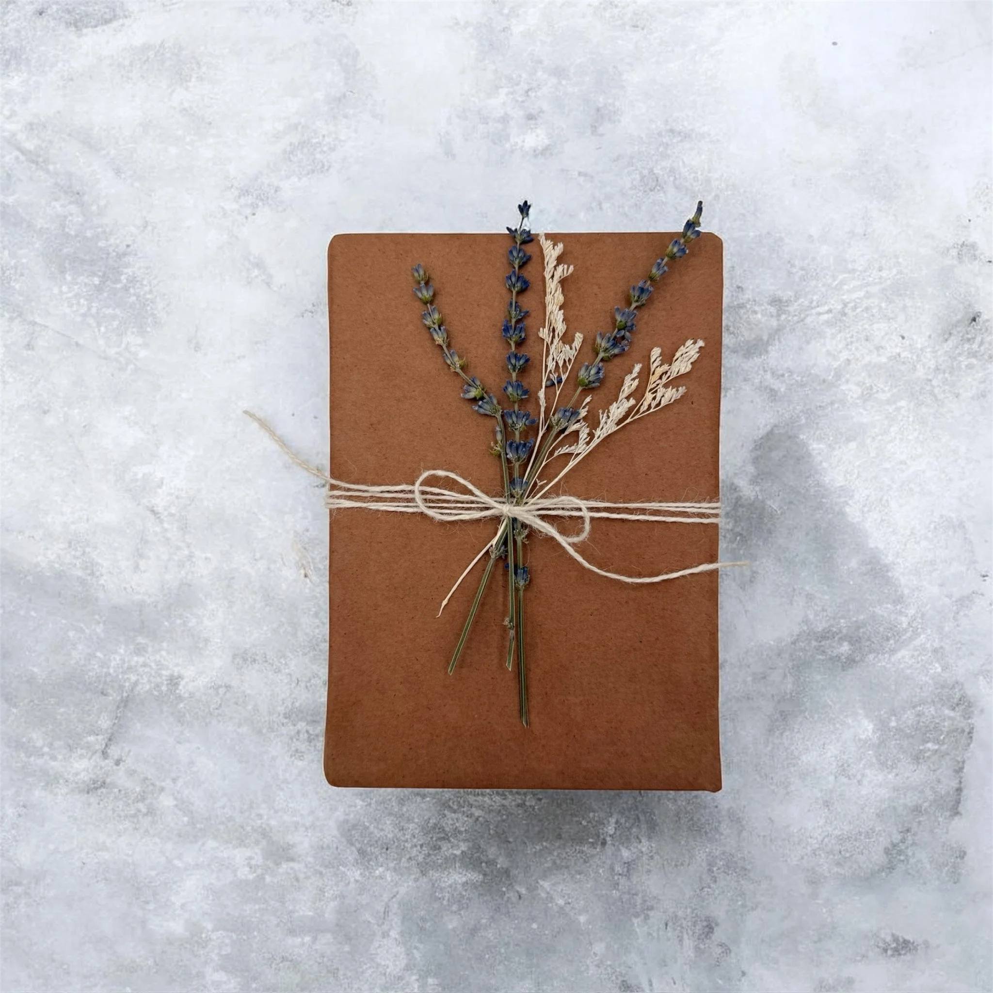 A brown wrapped gift box decorated with lavender sprigs tied with a piece of string on a light, textured background.
