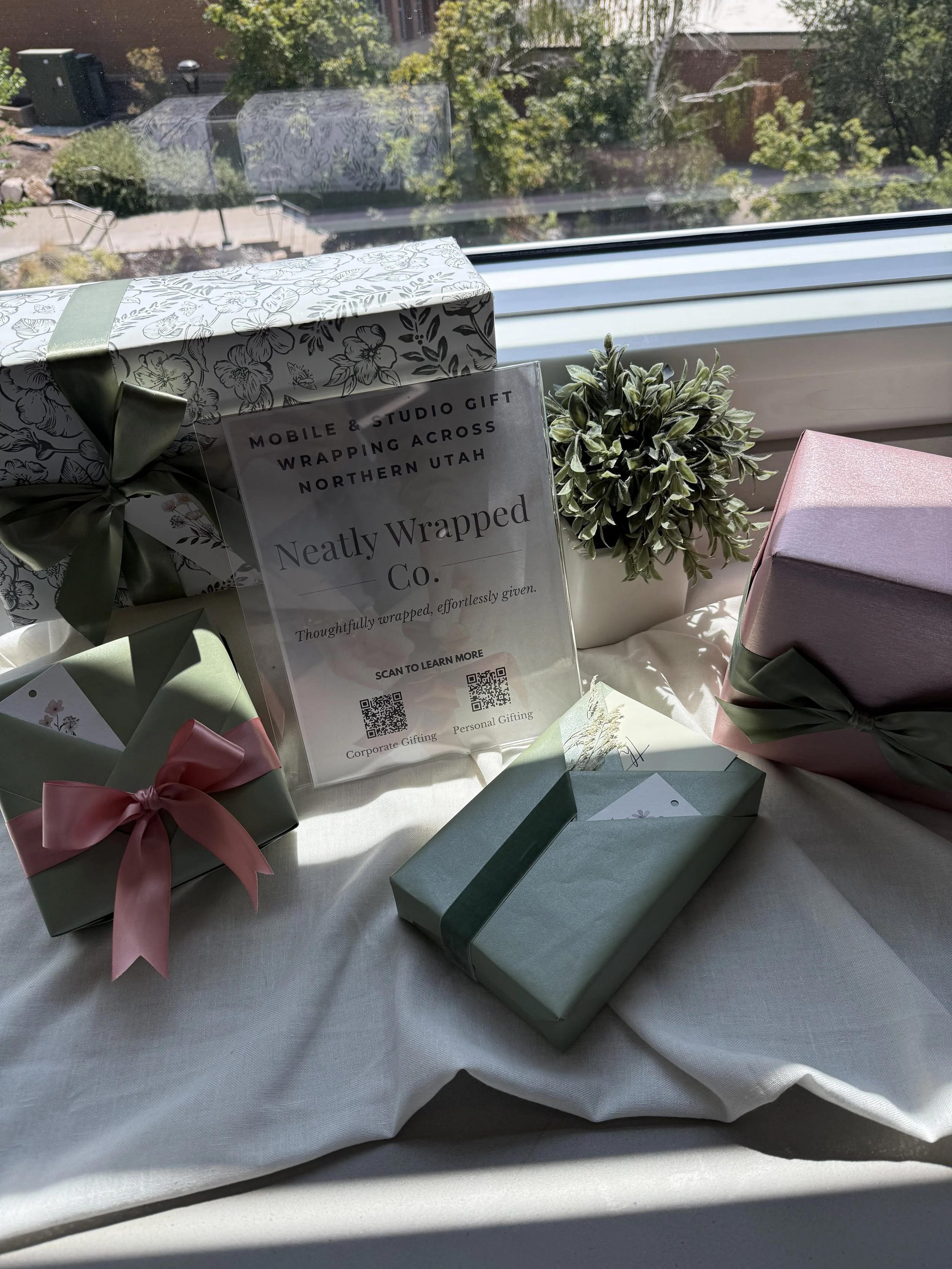 Various wrapped gifts with ribbons, a potted plant, and a sign promoting mobile and studio gift wrapping service on a window sill.