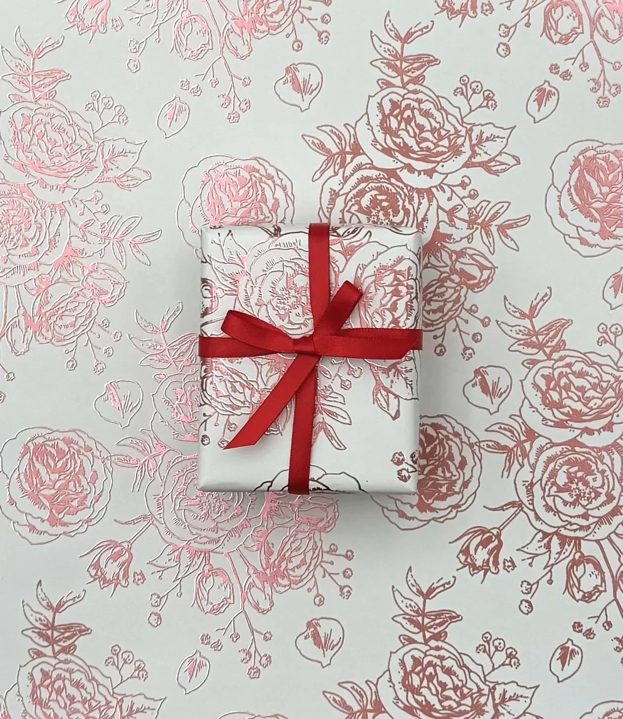 A small wrapped gift box with white wrapping paper featuring pink floral designs and tied with a red ribbon, placed on a matching pink floral patterned background.
