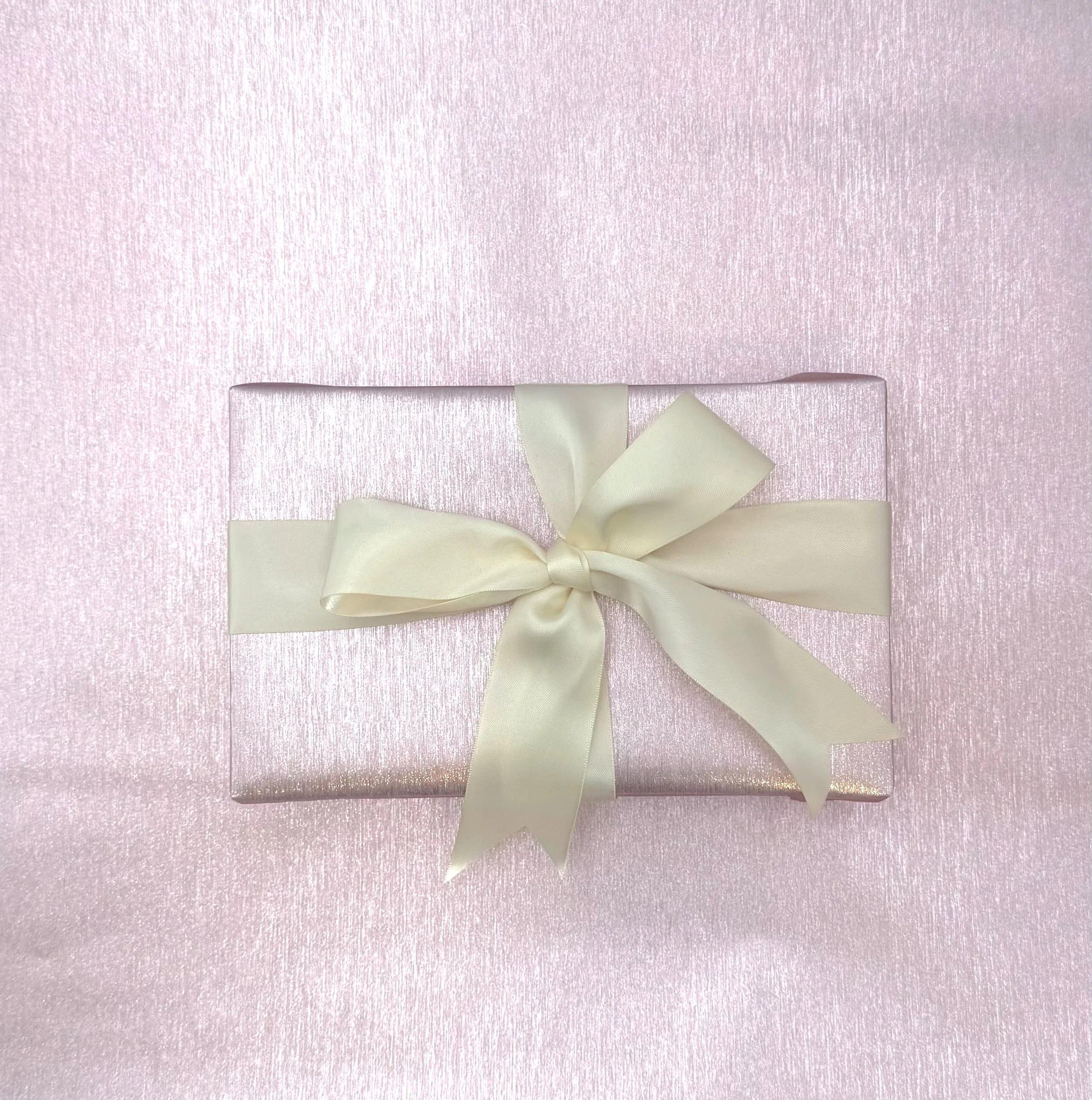 A pink textured gift box with a shiny finish, decorated with a cream satin ribbon tied into a bow, placed on a pink surface.