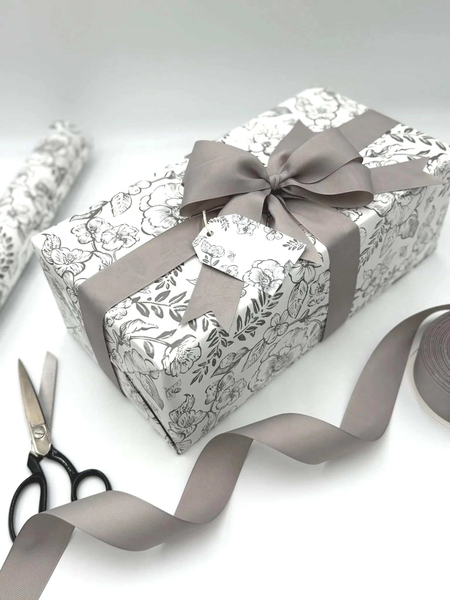 Decorative gift box wrapped in floral patterned paper with a gray ribbon bow, flat scissors, and a roll of gray ribbon on a white background.