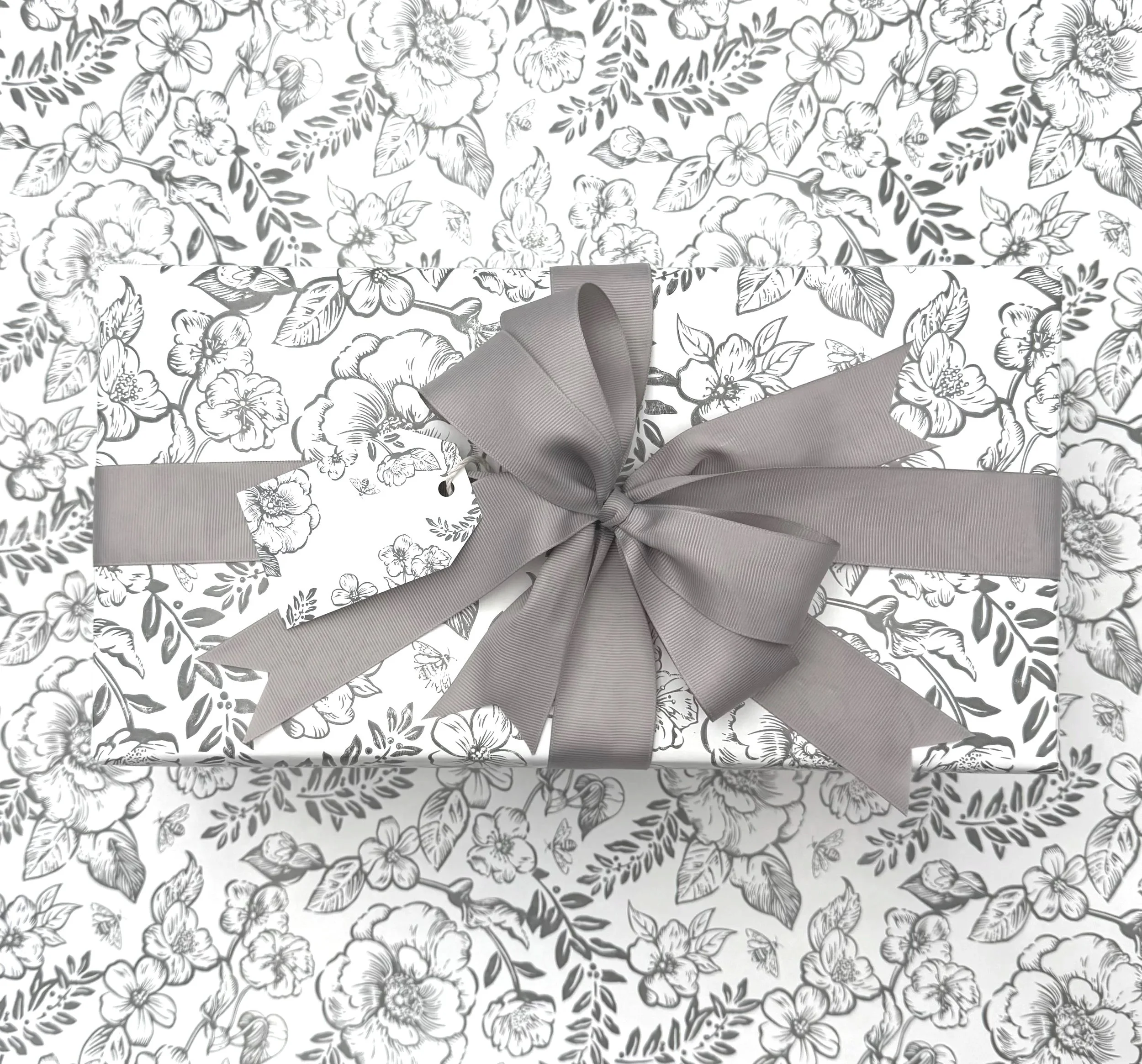 A rectangular gift wrapped in floral patterned paper with a gray ribbon and bow with a tag attached.