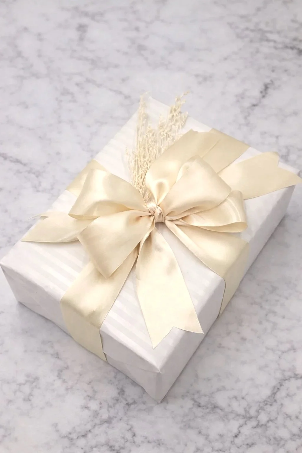 A gift wrapped in white paper with a gold satin ribbon tied in a bow, placed on a gray marble surface.