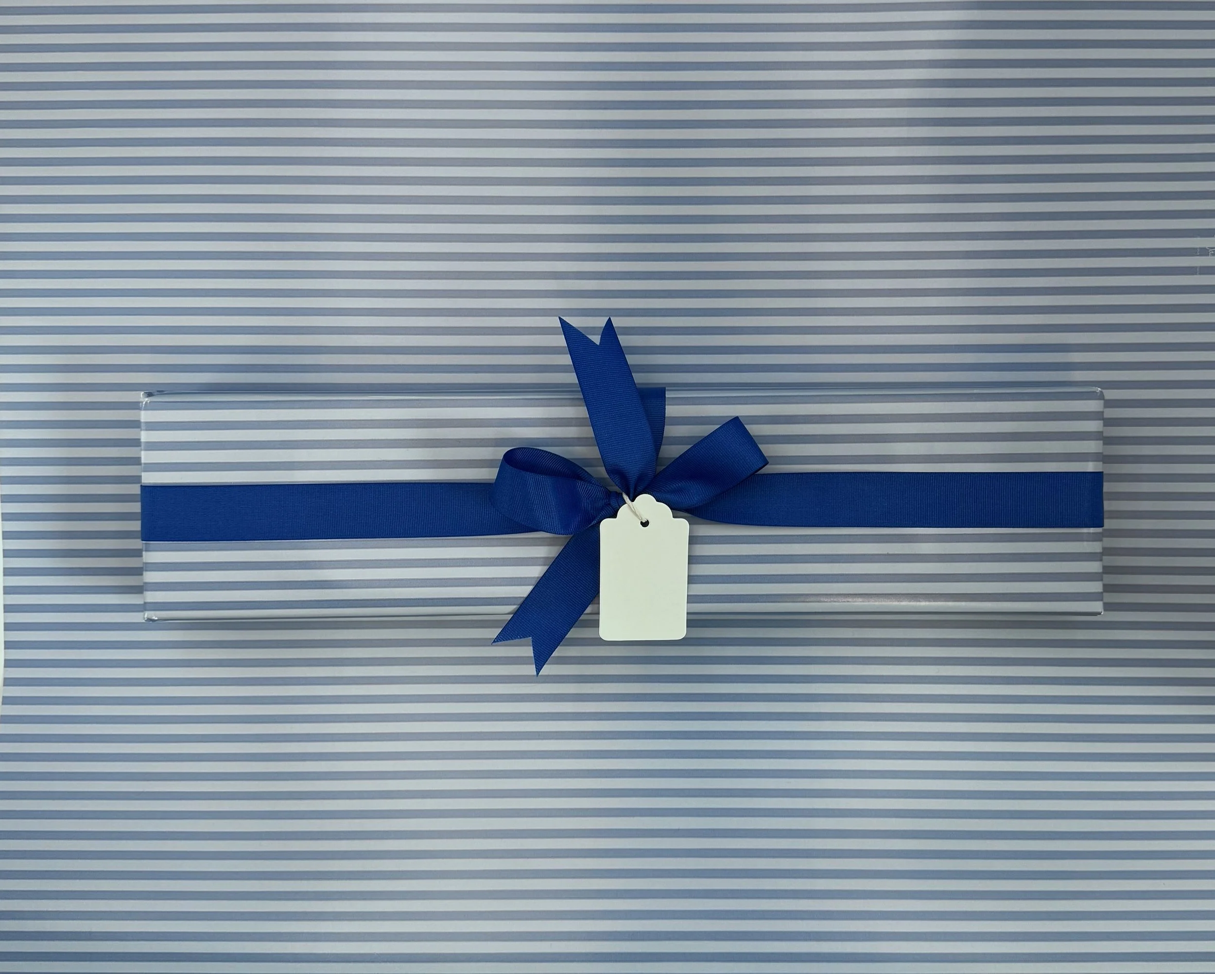 A rectangular gift box wrapped in white and silver striped paper, decorated with a blue ribbon tied in a bow, and a blank white gift tag attached.