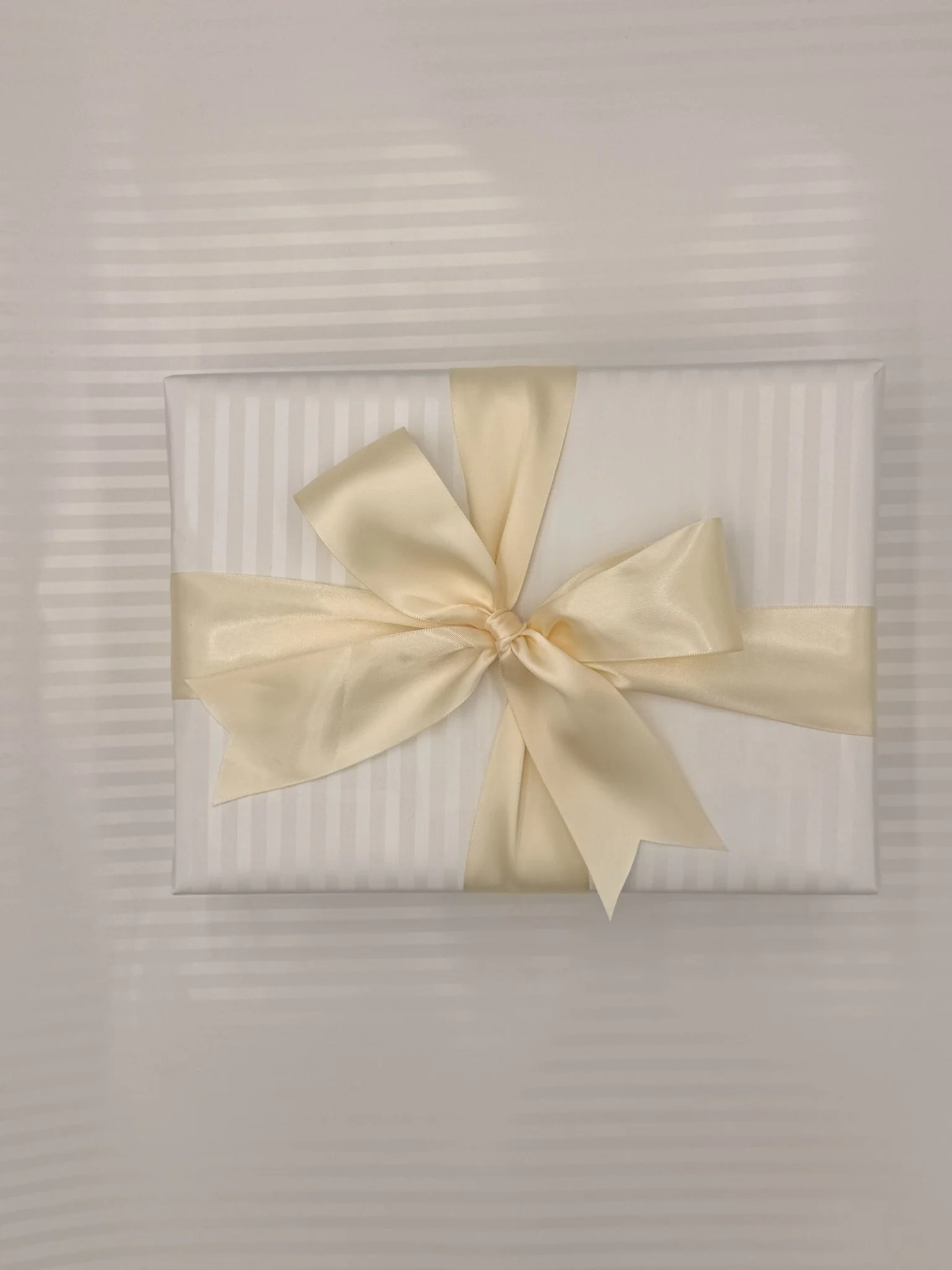 White gift box wrapped with a satin cream ribbon tied in a bow, resting on striped wrapping paper.