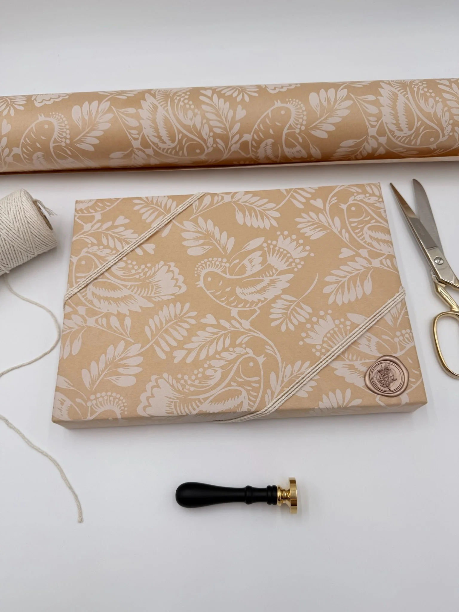 Wrapping paper with floral design, beige and white, with a wax seal, scissors, twine, and a wax stamp on a white surface.