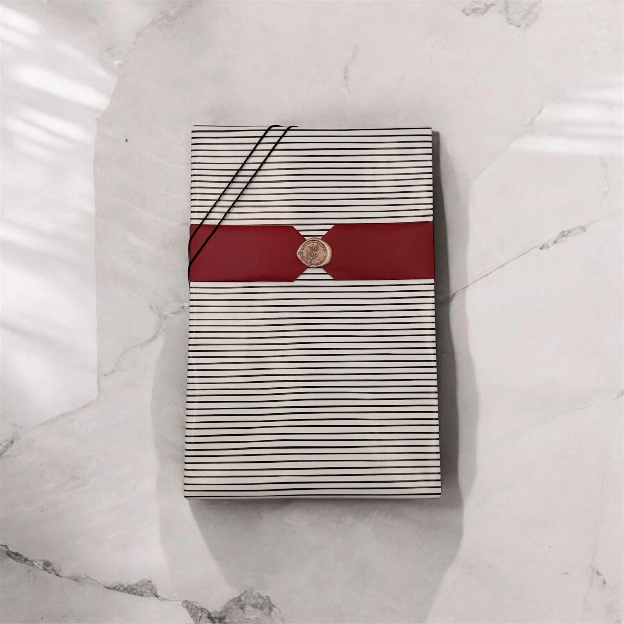 A notebook with black and white striped cover, red band with a wax seal, and two black strings on top, placed on a marble surface.