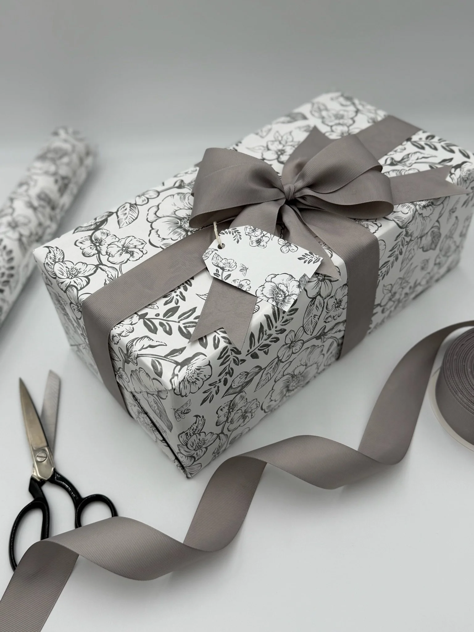 Decorative gift box wrapped in floral patterned paper with a gray ribbon bow, flat scissors, and a roll of gray ribbon on a white background.