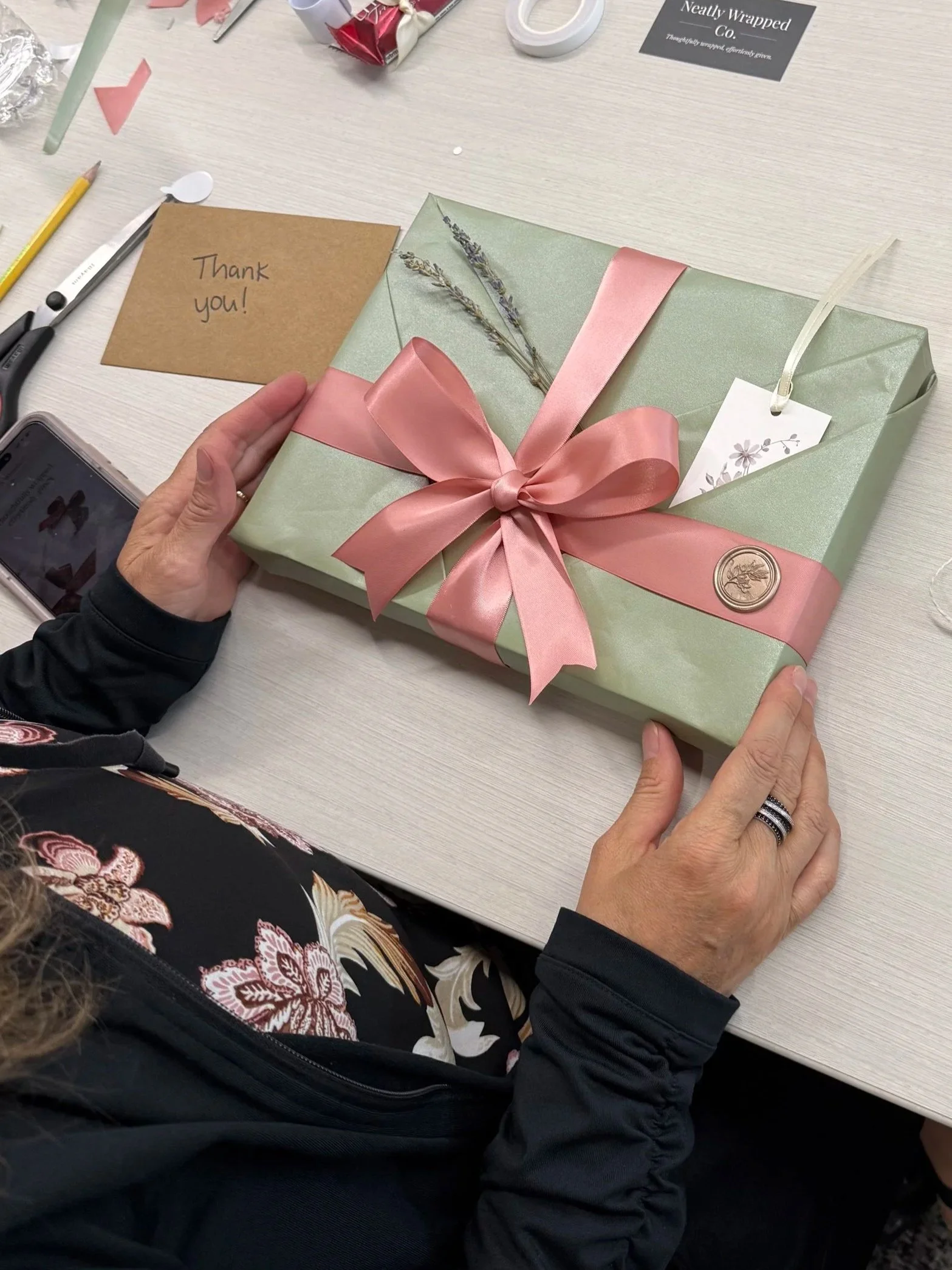 A person holding a wrapped gift box with a pink ribbon. On the table, there is a note card with 'Thank you!' written on it, scissors, and other gift wrapping supplies.