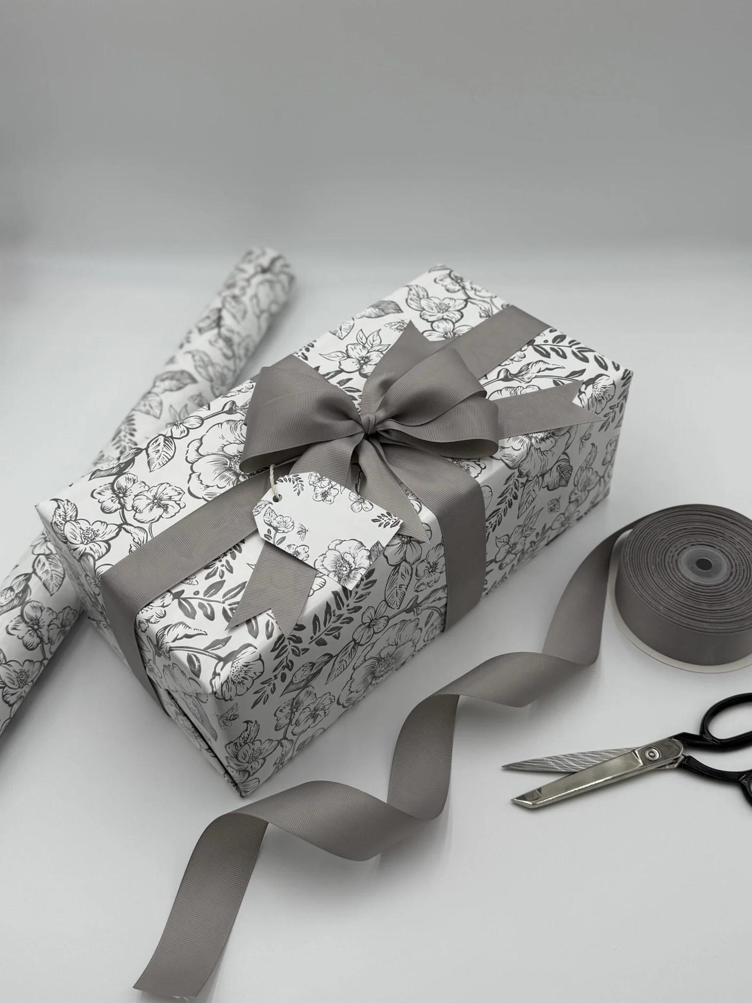 A gift box with black and white floral wrapping and a gray ribbon bow, surrounded by a roll of gray ribbon, scissors, and wrapping paper on a white surface.