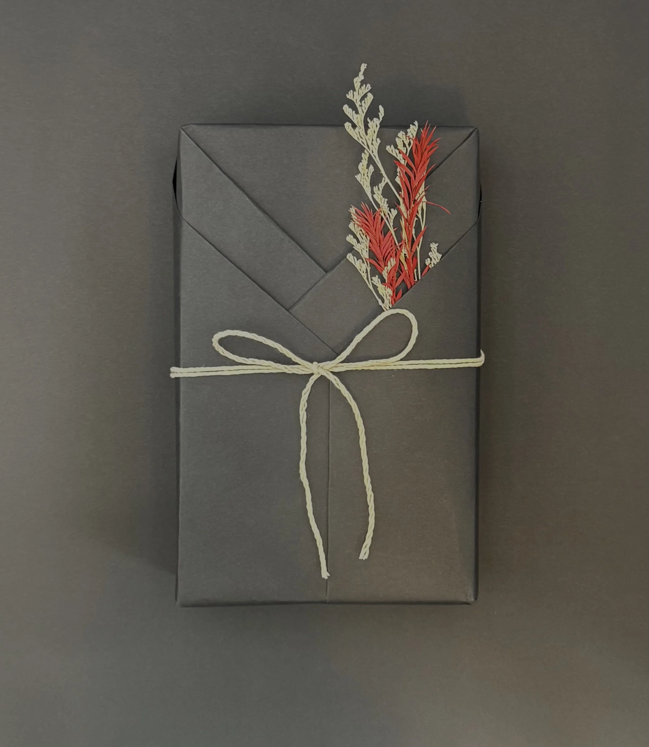 Gray gift box wrapped with white string, decorated with dried red and beige flowers, on a dark gray background.